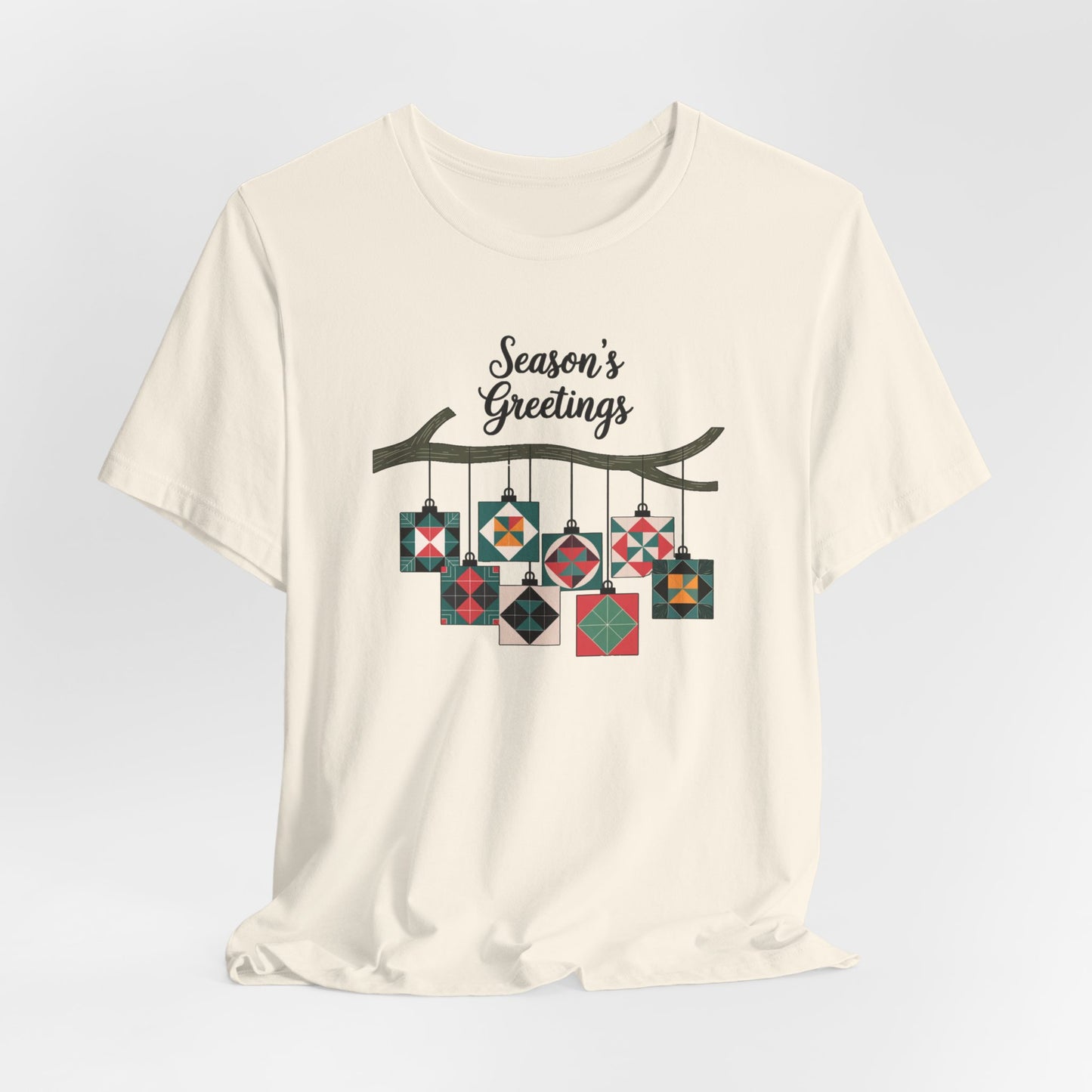 A Natural christmas quilting t-shirt featuring quilt block ornaments hanging from a branch, featuring the phrase "Season's Greetings" above. The ornaments showcase various quilting patterns to highlight the holiday quilting theme.