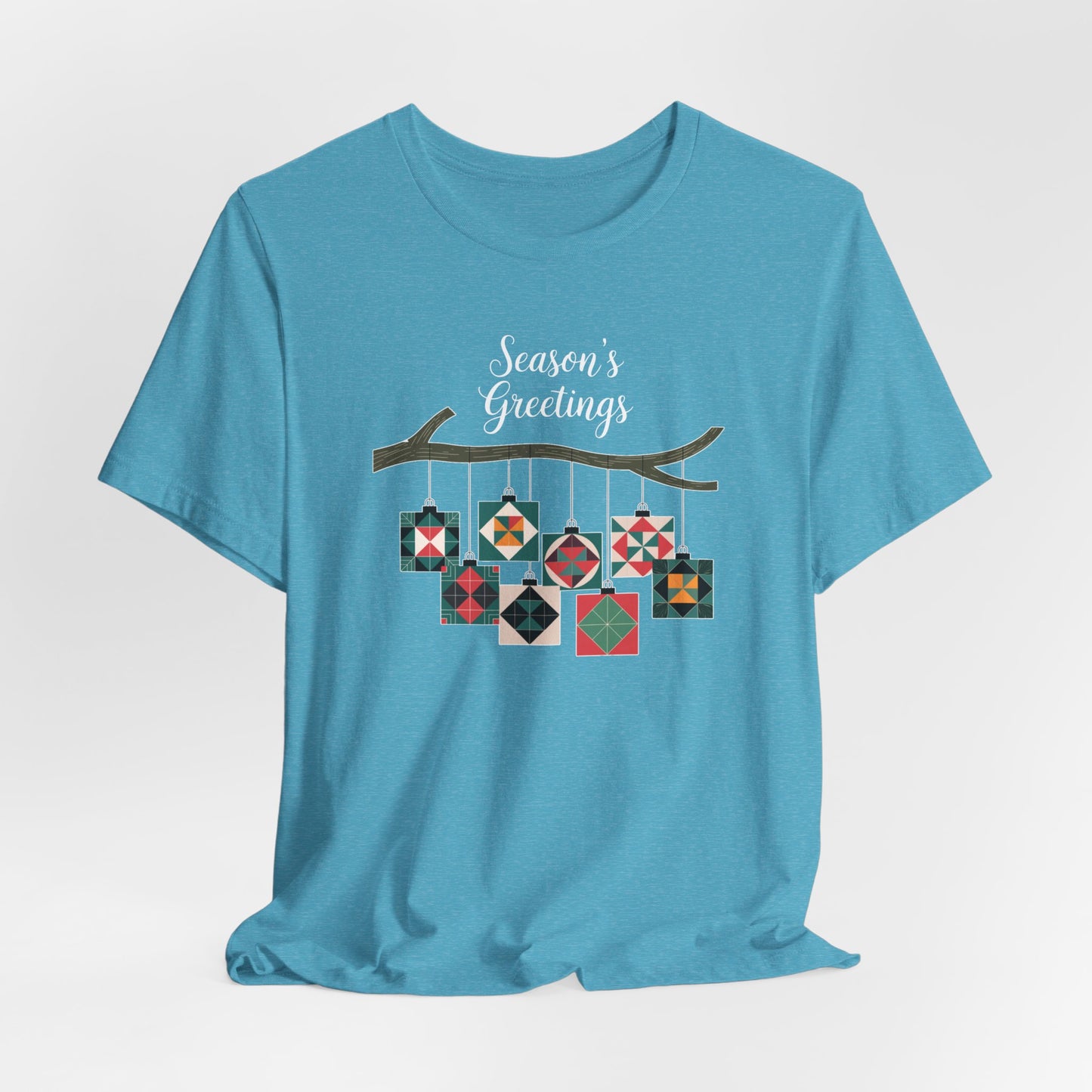 A Heather Aqua christmas quilting t-shirt featuring quilt block ornaments hanging from a branch, featuring the phrase "Season's Greetings" above. The ornaments showcase various quilting patterns to highlight the holiday quilting theme.