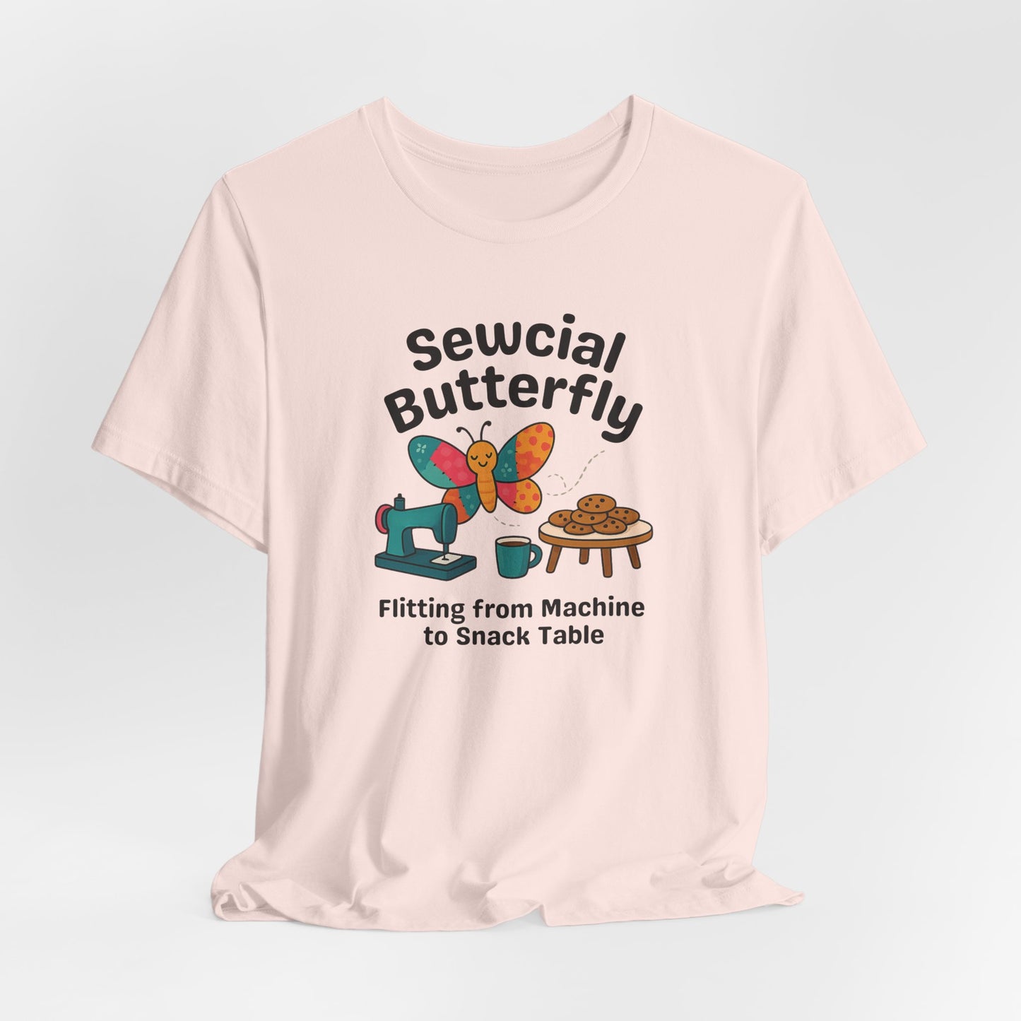A A Soft Pink t-shirt with a colorful design showing a patchwork butterfly flying between a sewing machine and a table of cookies. Text reads “Sewcial Butterfly – Flitting from Machine to Snack Table.” Funny gift for quilters