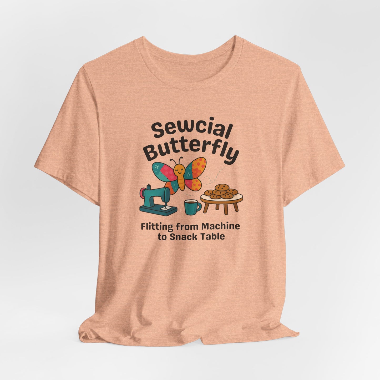 A A Heather Peach t-shirt with a colorful design showing a patchwork butterfly flying between a sewing machine and a table of cookies. Text reads “Sewcial Butterfly – Flitting from Machine to Snack Table.” Funny gift for quilters