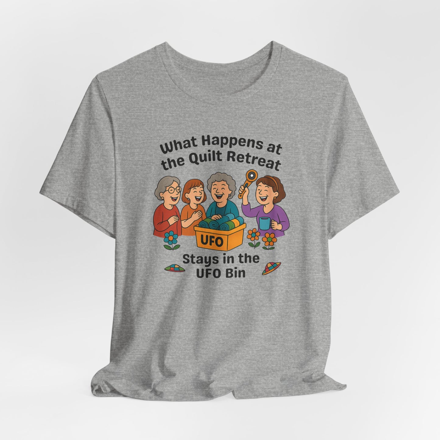 A An Athletic Heather t-shirt featuring a cartoon design of four laughing women at a quilt retreat with a bin labeled “UFO” full of fabric. Surrounded by patchwork flying saucers and flowers. Text reads “What happens at the quilt retreat stays in the UFO bin.”