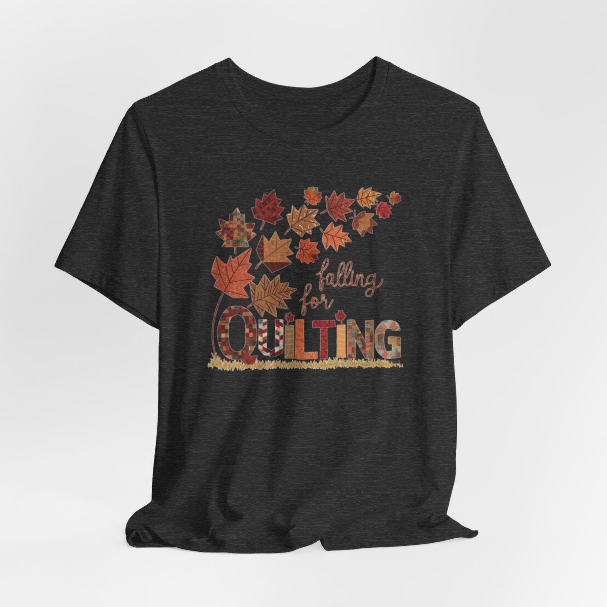 A Dark Grey Heather thanksgiving quilting t-shirt featuring autumn leaves with various quilting patterns inside. Text reads "falling for QUILTING" with letters in patchwork style. Warm fall colors and quilting motifs combine in a seasonal design.