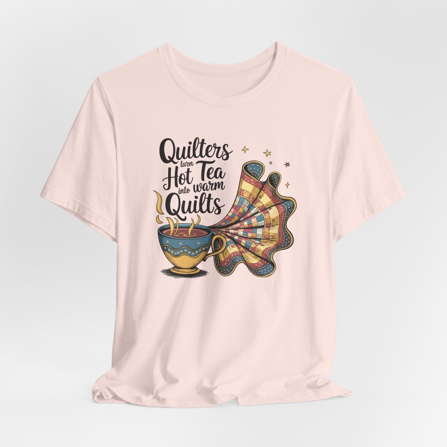 A Soft Pink t-shirt with a whimsical design of a teacup transforming into a patchwork quilt, accompanied by the phrase 'Quilters turn hot tea into warm quilts.' Ideal for quilters and tea lovers
