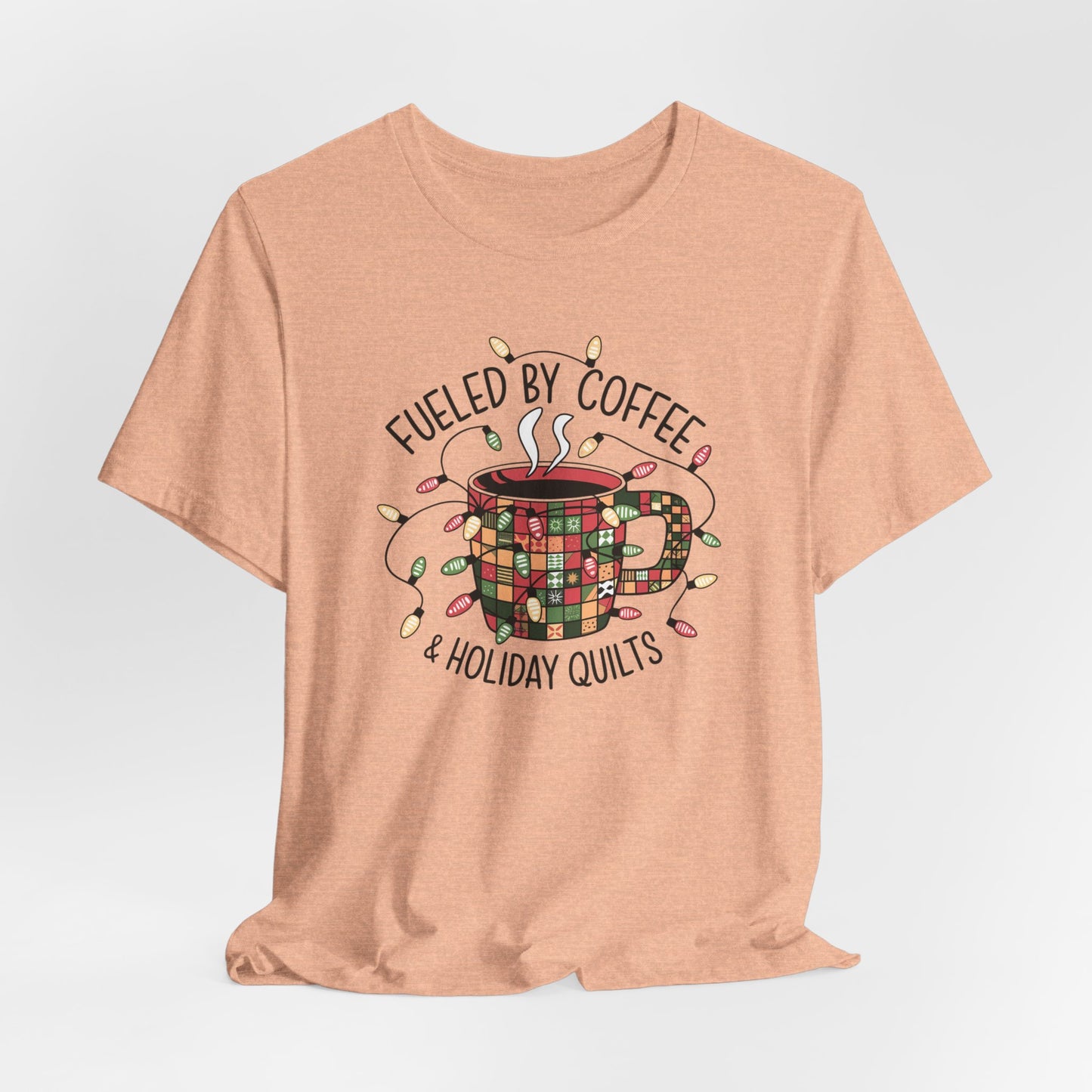 A Heather Peach christmas quilting t-shirt featuring a cozy coffee mug covered in patchwork quilt patterns, wrapped in festive holiday lights, with the phrase "Fueled by Coffee & Holiday Quilts" surrounding the design.