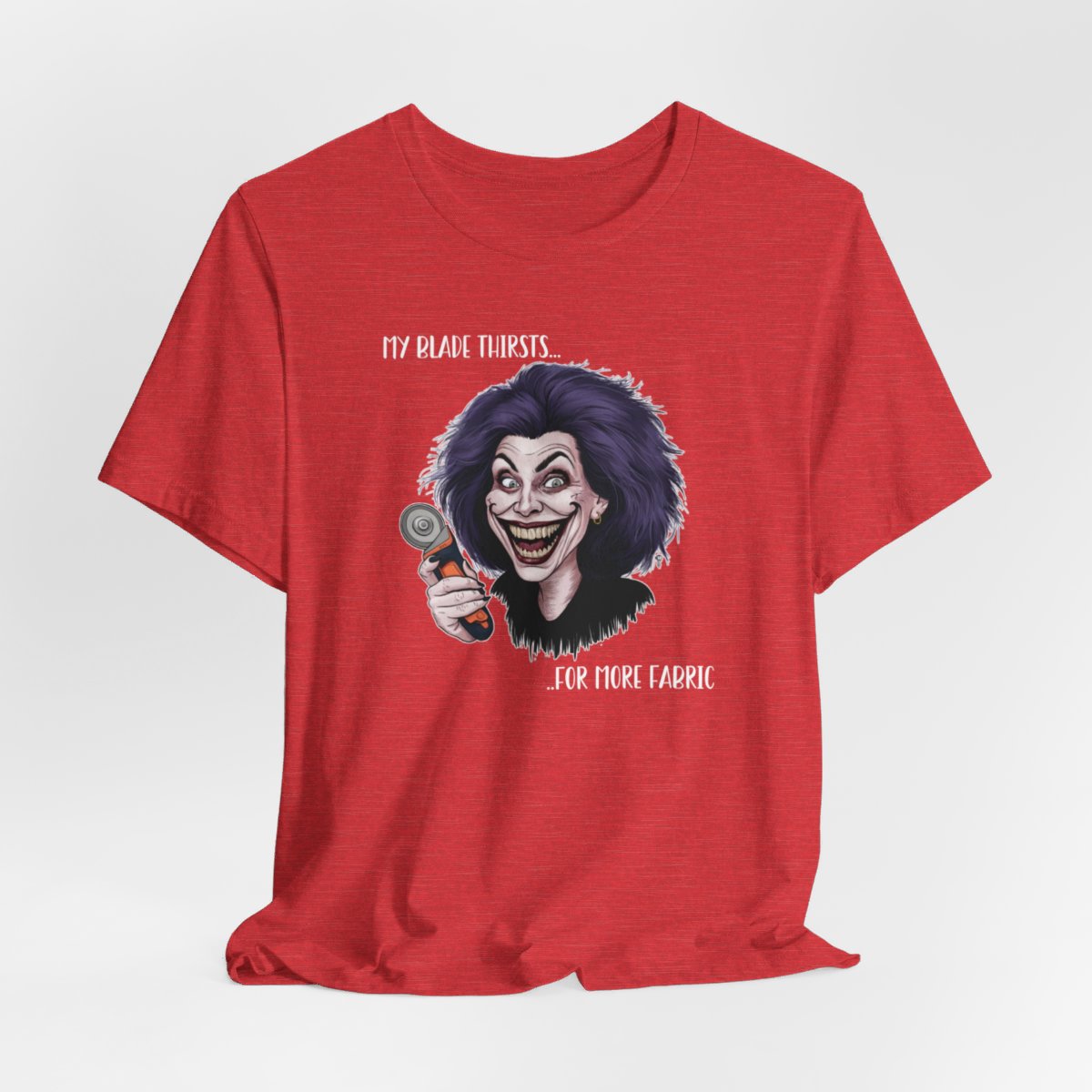 A Heather Red T-shirt showing a cartoonish character with wild purple hair and an exaggerated grin, holding a rotary cutter. Text reads "My blade thirsts... for more fabric", humorously depicting a quilter's endless desire for new materials.