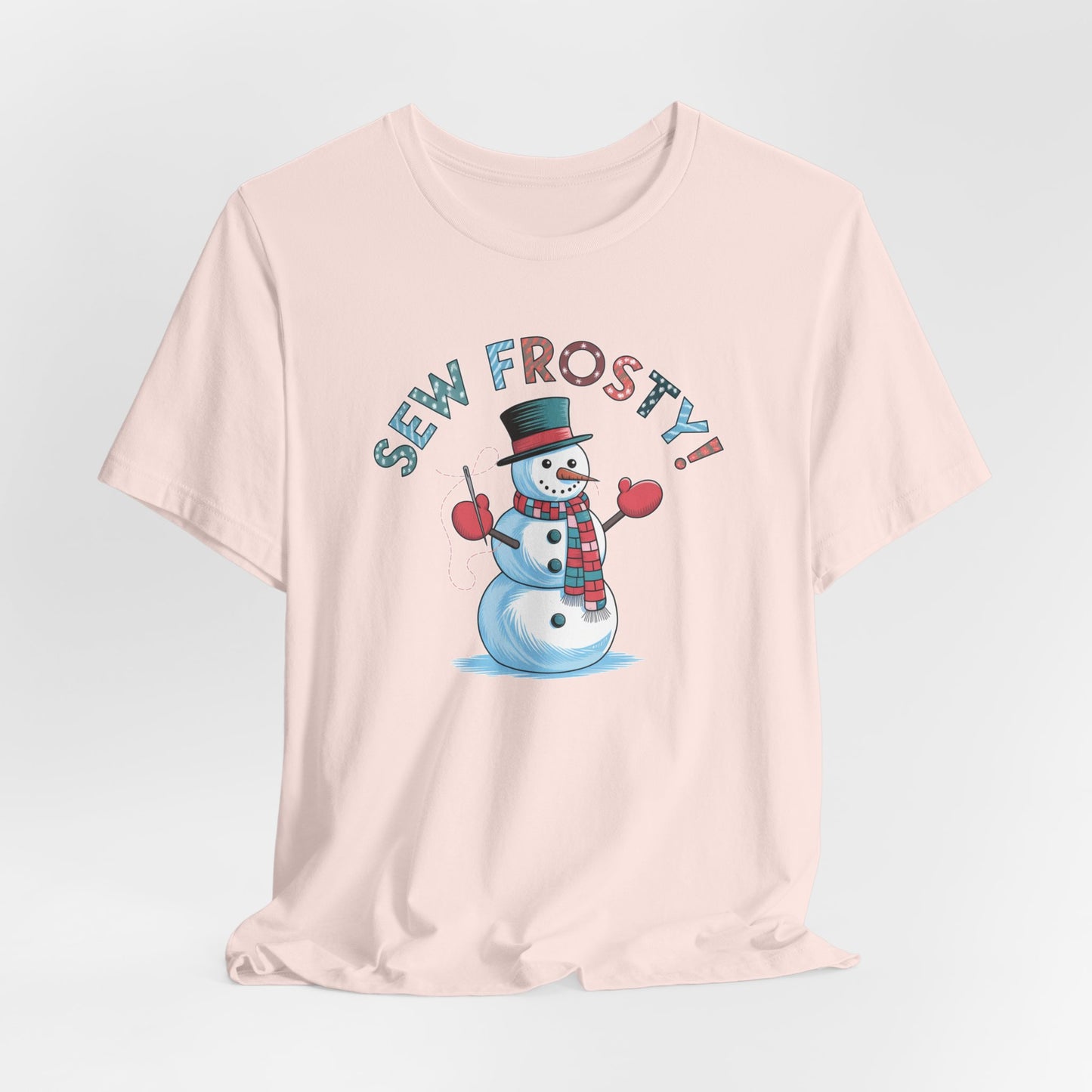 A Soft Pink christmas quilting t-shirt featuring cheerful snowman design holding a sewing needle, with the phrase "Sew Frosty" in playful, winter-themed lettering. The snowman is wearing a scarf and mittens, ready for holiday sewing fun.