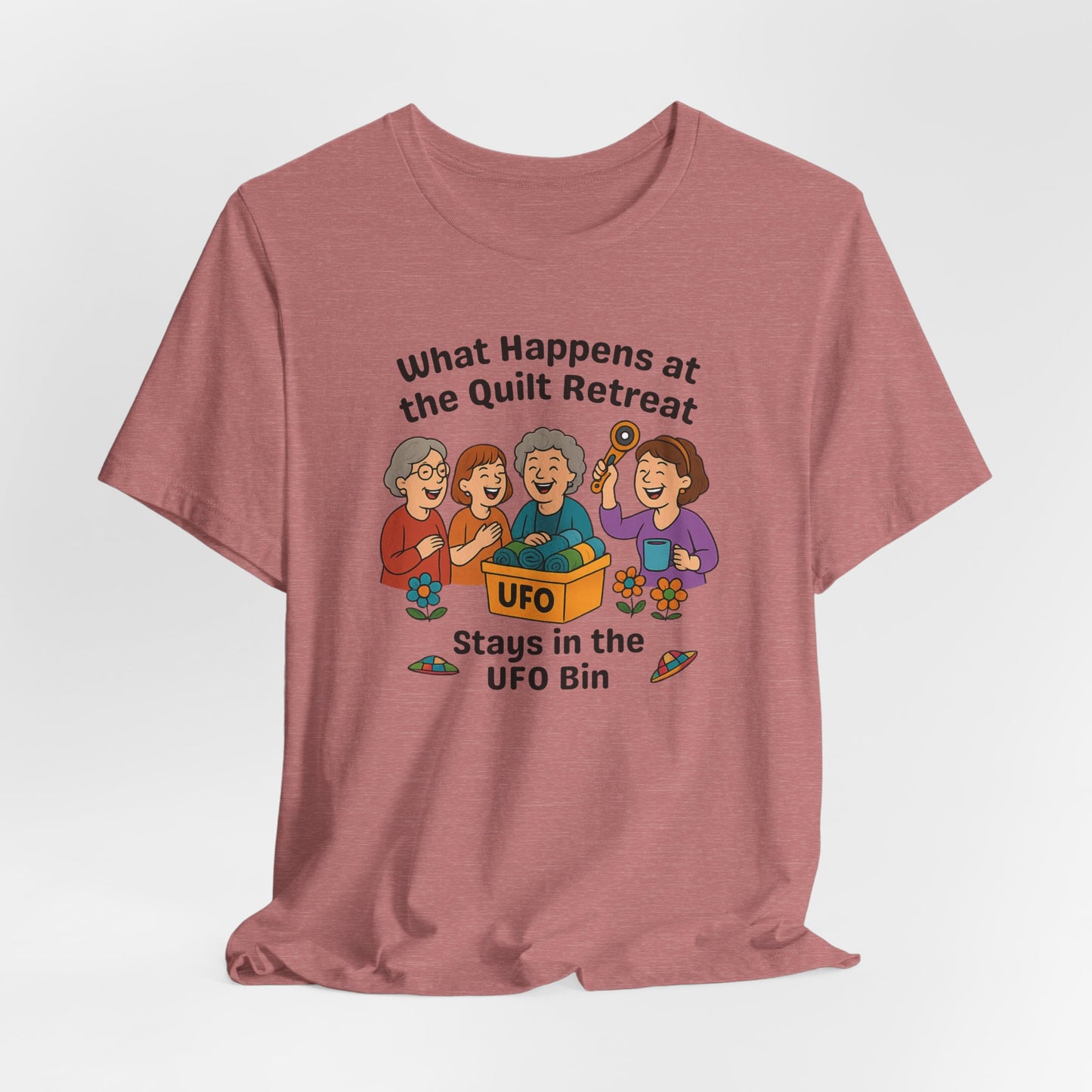 A A Heather Mauve t-shirt featuring a cartoon design of four laughing women at a quilt retreat with a bin labeled “UFO” full of fabric. Surrounded by patchwork flying saucers and flowers. Text reads “What happens at the quilt retreat stays in the UFO bin.”