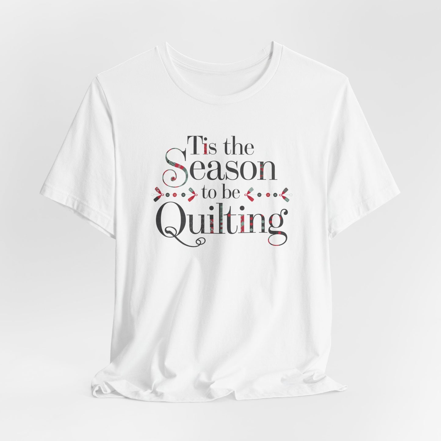 A White festive t-shirt featuring the phrase "'Tis the Season to be Quilting," with playful sewing-inspired elements like needles, buttons, and fabric patterns integrated into the typography.