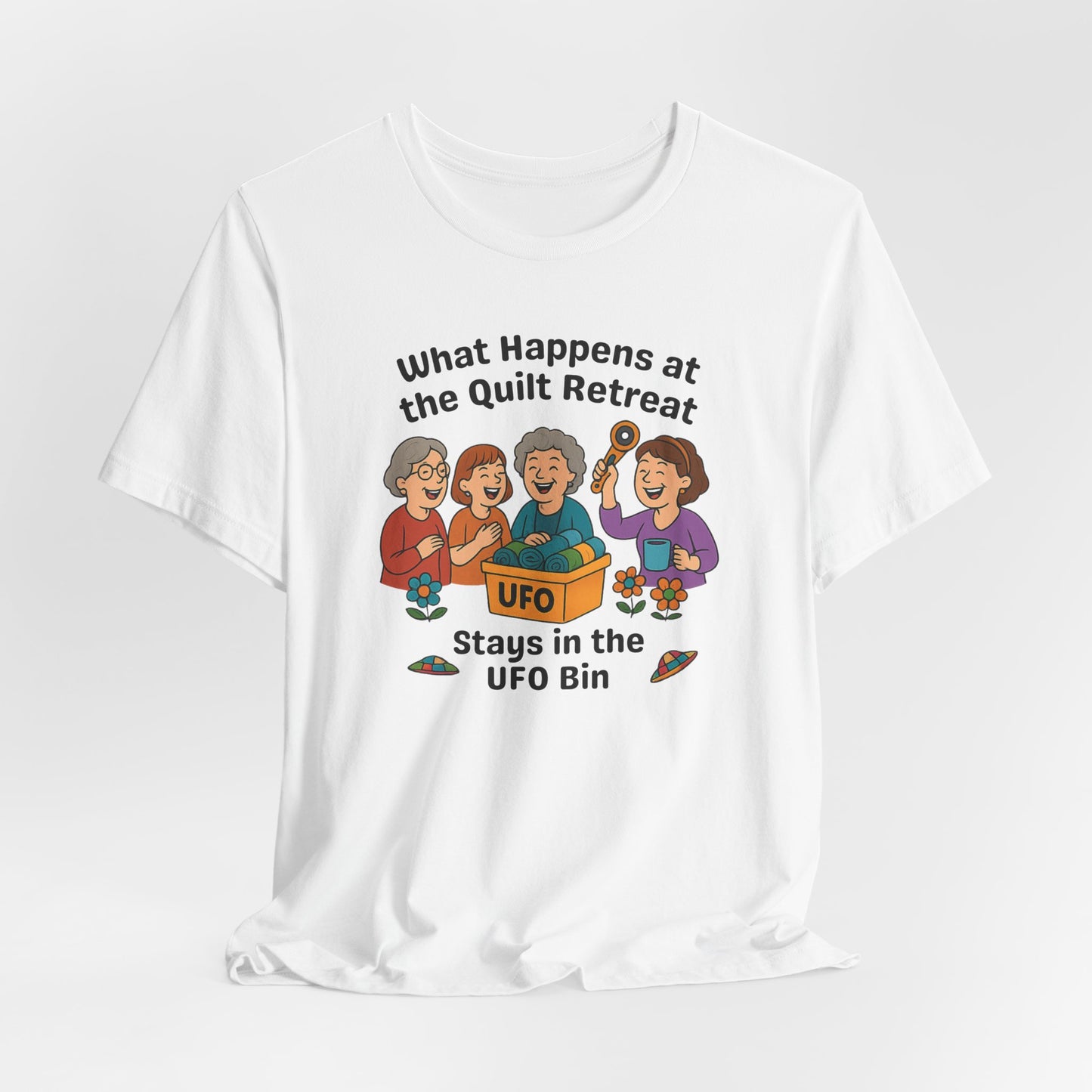 A A White t-shirt featuring a cartoon design of four laughing women at a quilt retreat with a bin labeled “UFO” full of fabric. Surrounded by patchwork flying saucers and flowers. Text reads “What happens at the quilt retreat stays in the UFO bin.”