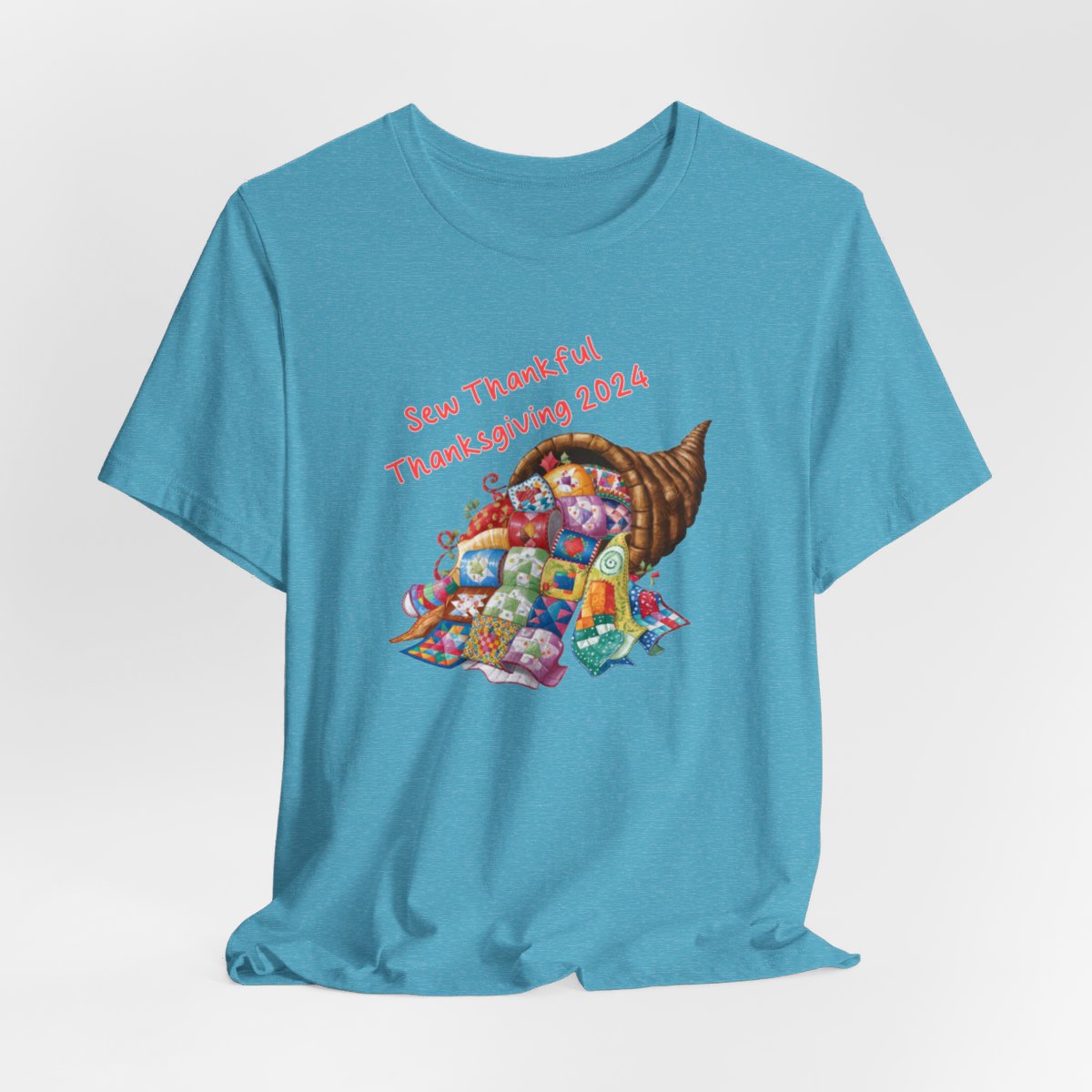 A Heather Aqua thanksgiving quilting t-shirt featuring a colorful quilted cornucopia overflowing with various quilt patterns and designs. Text reads "Sew Thankful Thanksgiving 2024" in red. Vibrant illustration combines quilting motifs with Thanksgiving imagery.