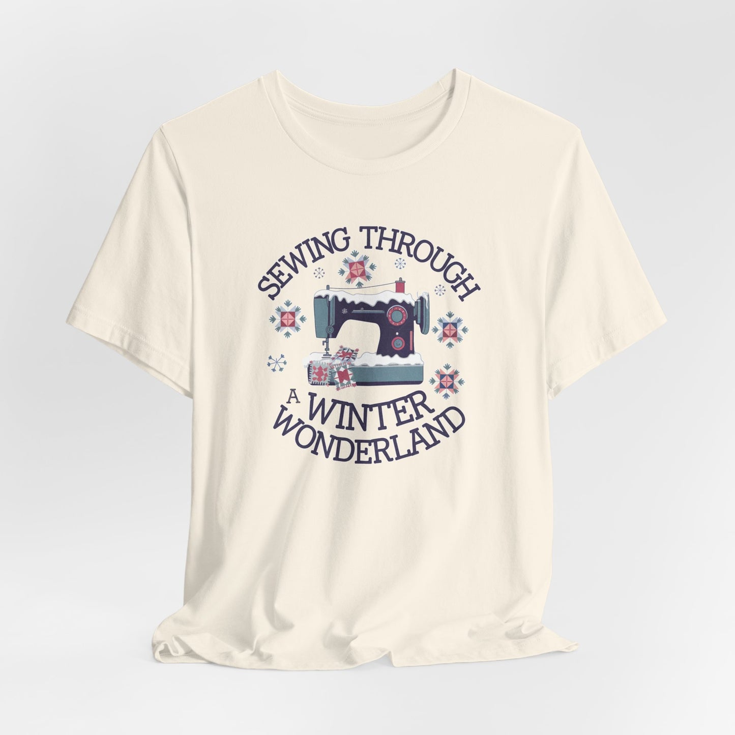 A Natural winter quilting t-shirt featuring a sewing machine covered in snow, surrounded by quilted snowflakes, with the phrase "Sewing Through a Winter Wonderland" above and below the image.