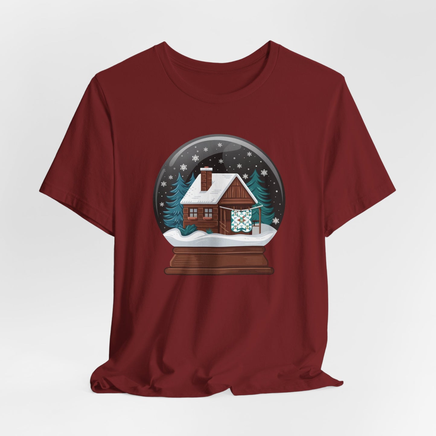 A Cardinal christmas quilting t-shirt featuring a cozy log cabin covered in snow, with a quilt hanging outside, set inside a snow globe with swirling snowflakes, blending quilting and holiday warmth.