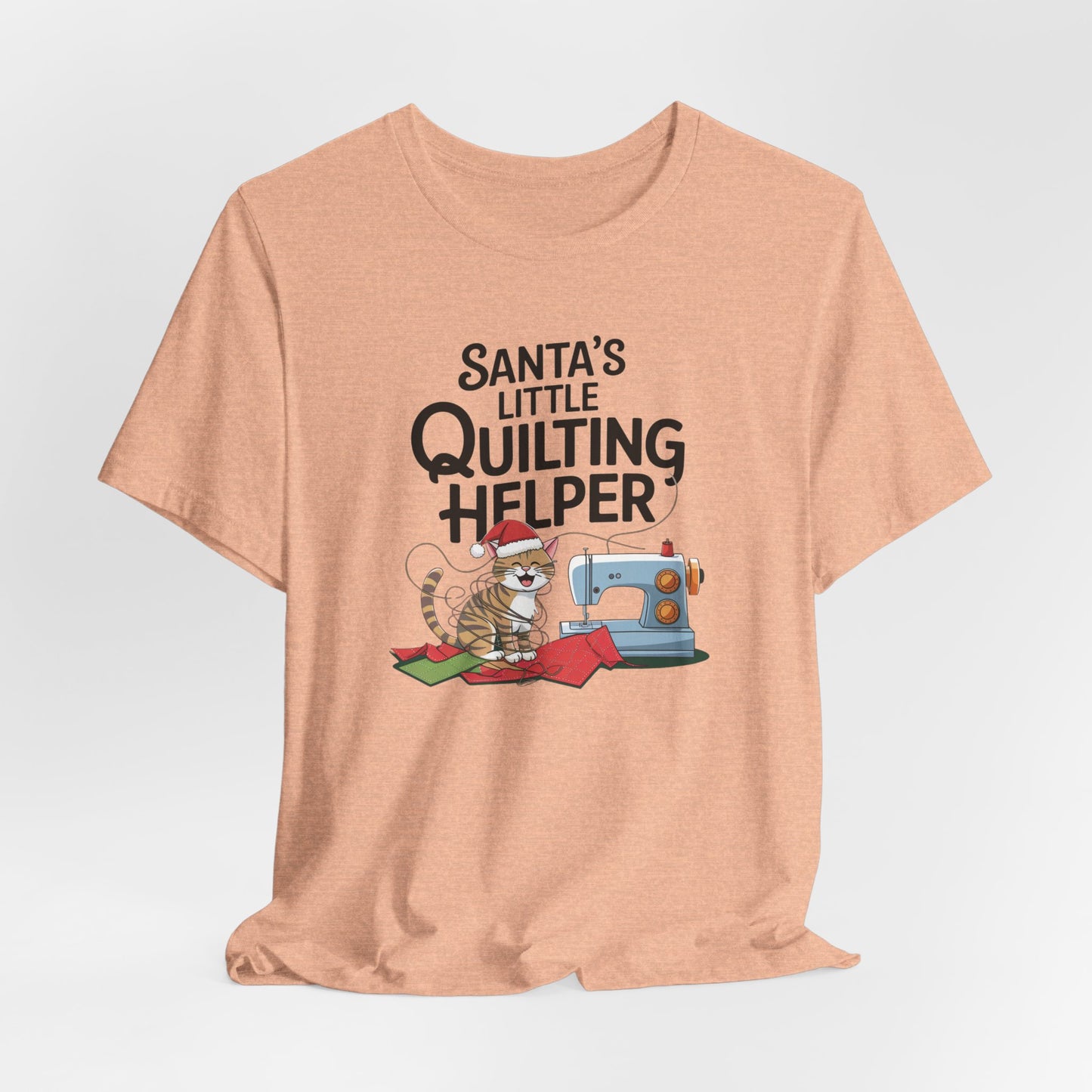 A Heather Peach christmas quilting t-shirt featuring a playful cat wearing a Santa hat is tangled in thread next to a sewing machine, with the phrase "Santa's Little Quilting Helper" above, combining quilting and holiday humor.