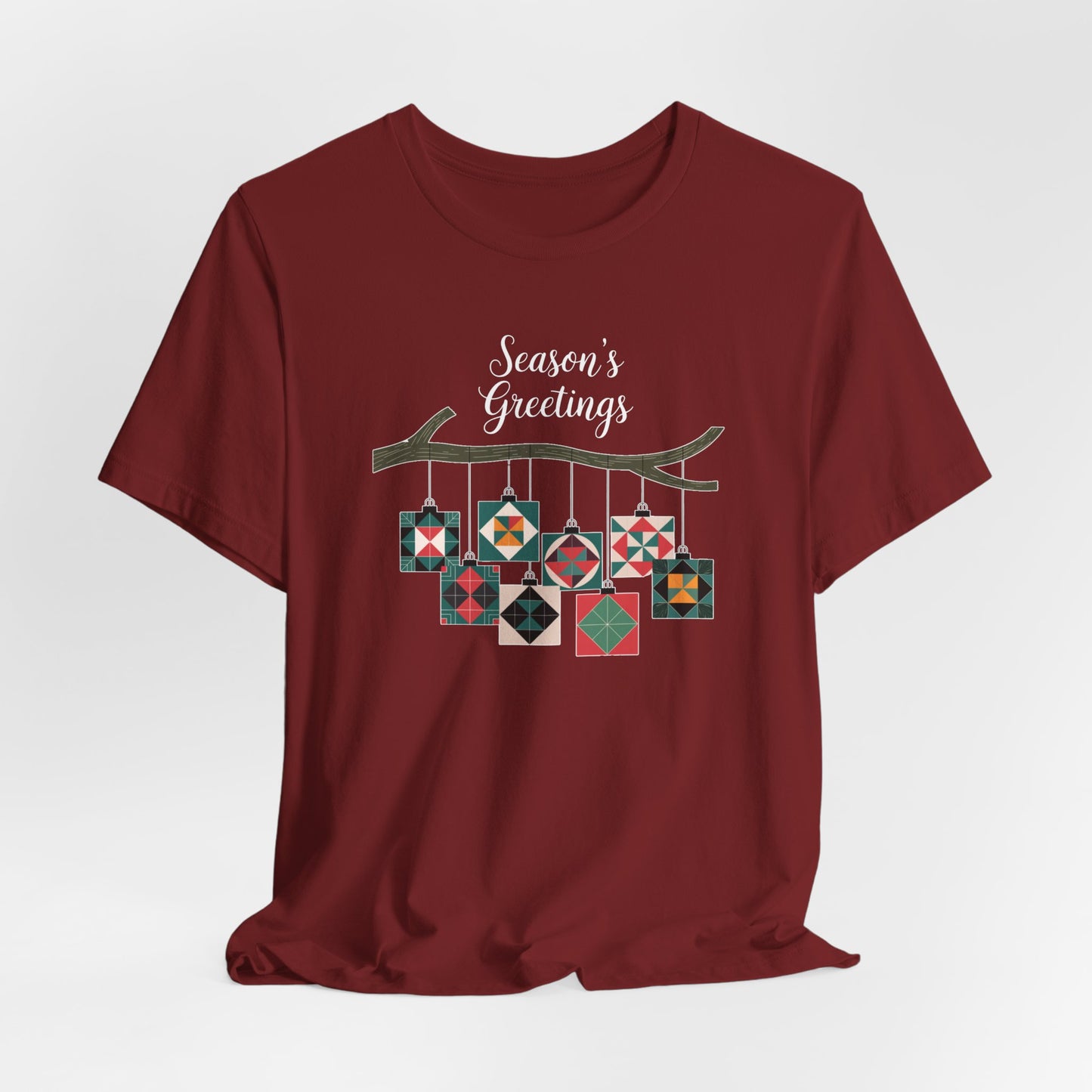 A Cardinal christmas quilting t-shirt featuring quilt block ornaments hanging from a branch, featuring the phrase "Season's Greetings" above. The ornaments showcase various quilting patterns to highlight the holiday quilting theme.
