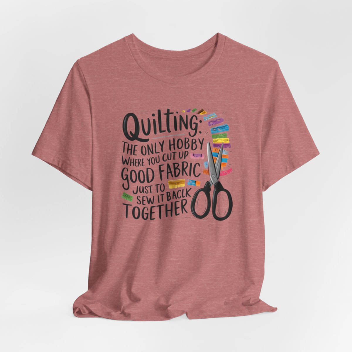 A Heather Mauve Quilting-themed T-Shirt with text "Quilting: The Only Hobby Where You Cut Up Good Fabric Just to Sew it Back Together" surrounded by colorful fabric scraps and scissors.