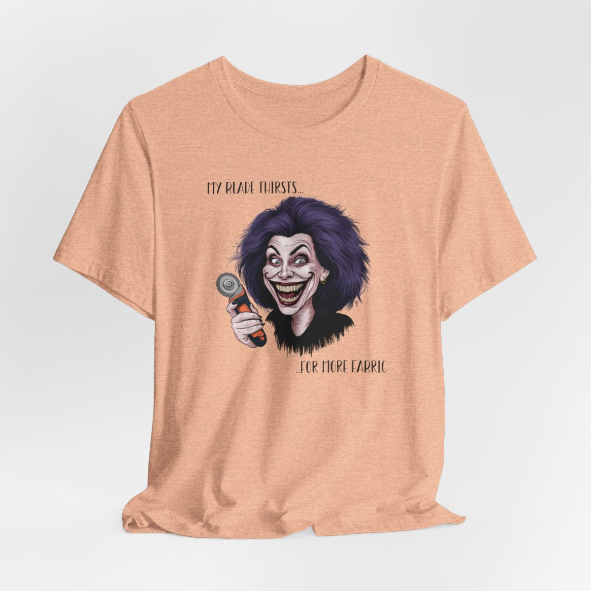 A Heather Peach T-shirt showing a cartoonish character with wild purple hair and an exaggerated grin, holding a rotary cutter. Text reads "My blade thirsts... for more fabric", humorously depicting a quilter's endless desire for new materials.