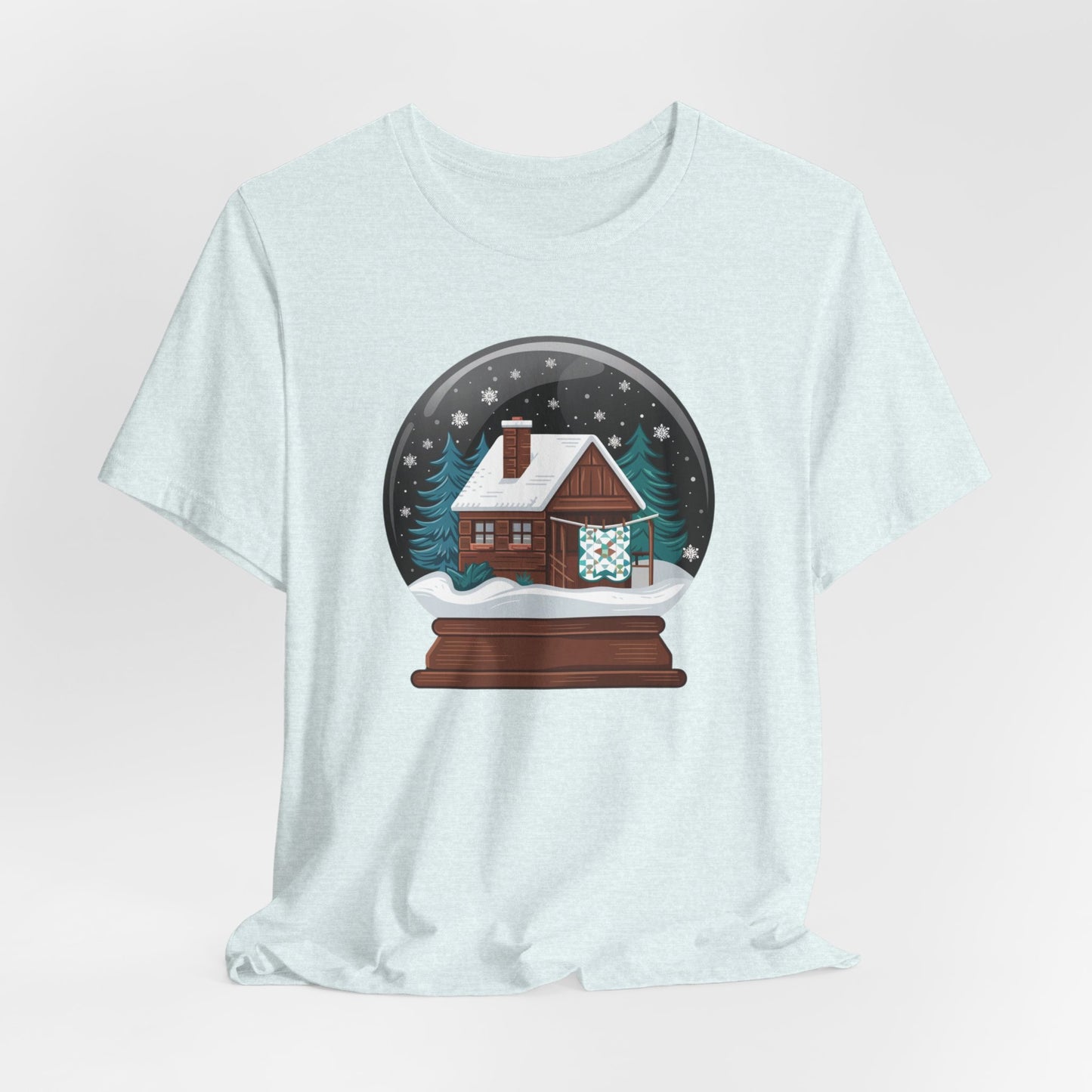 A Heather Ice Blue christmas quilting t-shirt featuring a cozy log cabin covered in snow, with a quilt hanging outside, set inside a snow globe with swirling snowflakes, blending quilting and holiday warmth.