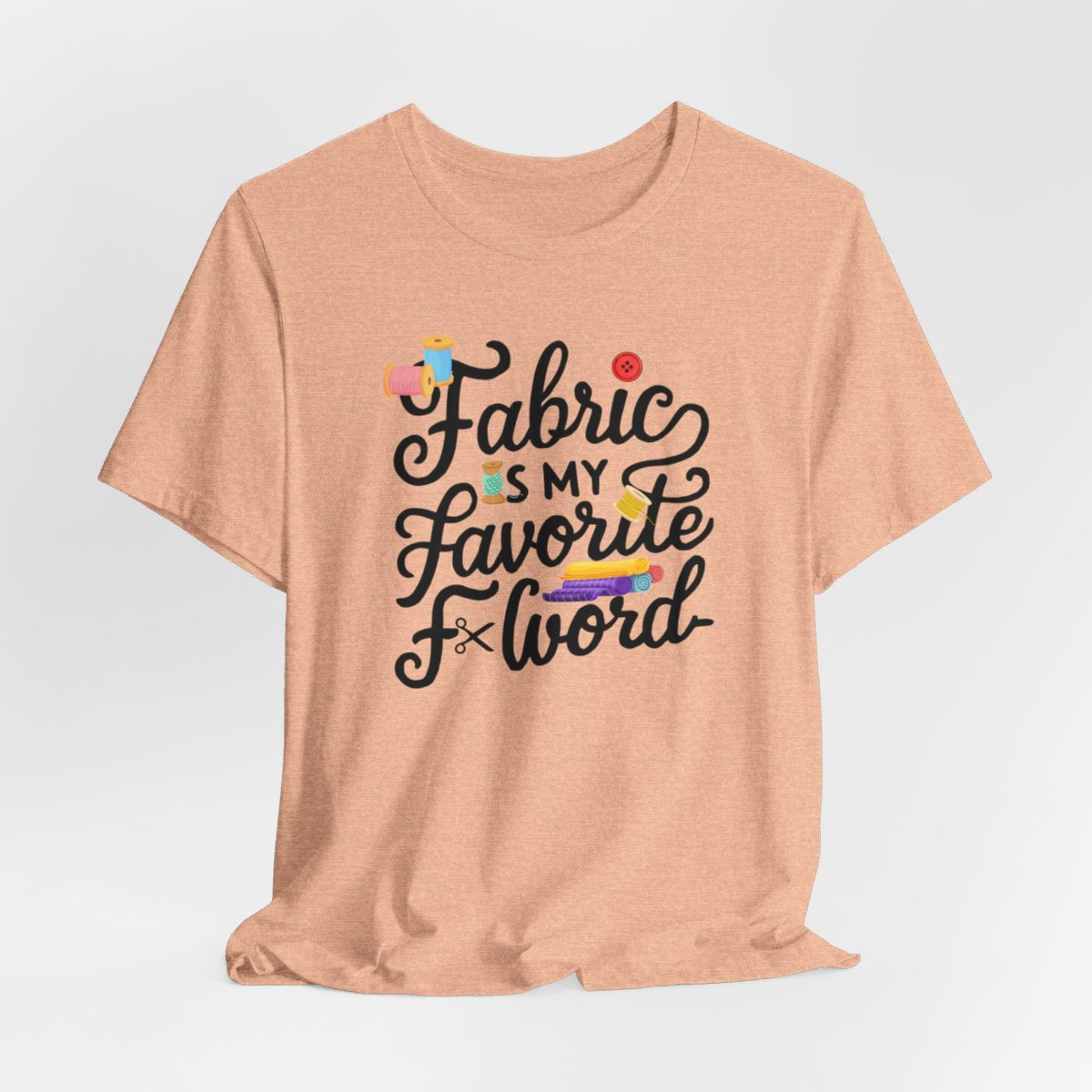 A Heather Peach T-Shirt with the phrase Fabric is My Favorite F-Word in a bold font with thread, buttons, and fabric bolts
