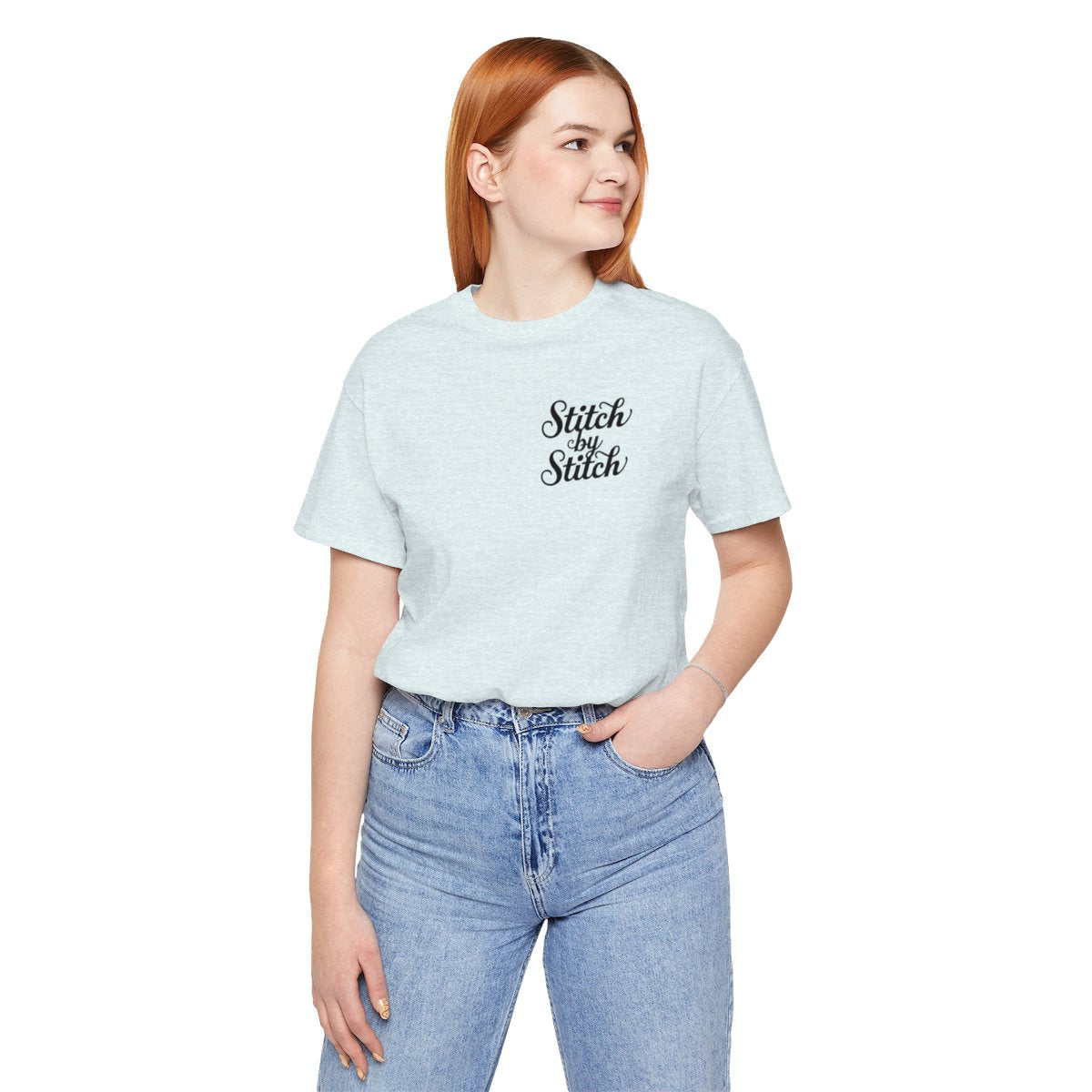 A Heather Ice Blue T-Shirt featuring the phrase "Stitch by Stitch". The word "Stitch" is written twice in a flowing script font, while "by" is centered between them in a smaller, sans-serif font. The design has a clean, minimalist aesthetic.