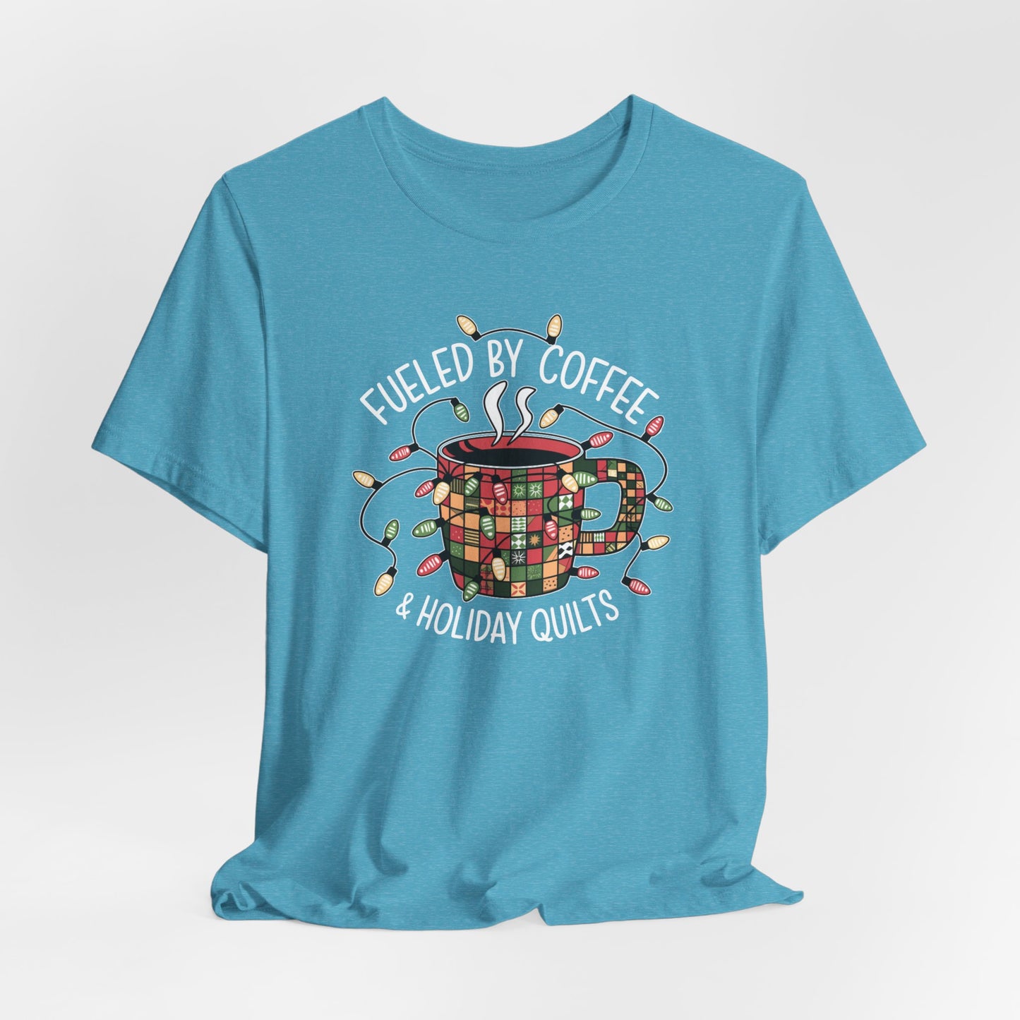A Heather Aqua christmas quilting t-shirt featuring a cozy coffee mug covered in patchwork quilt patterns, wrapped in festive holiday lights, with the phrase "Fueled by Coffee & Holiday Quilts" surrounding the design.