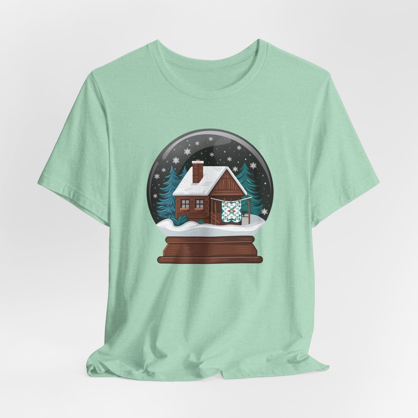 A Heather Mint christmas quilting t-shirt featuring a cozy log cabin covered in snow, with a quilt hanging outside, set inside a snow globe with swirling snowflakes, blending quilting and holiday warmth.