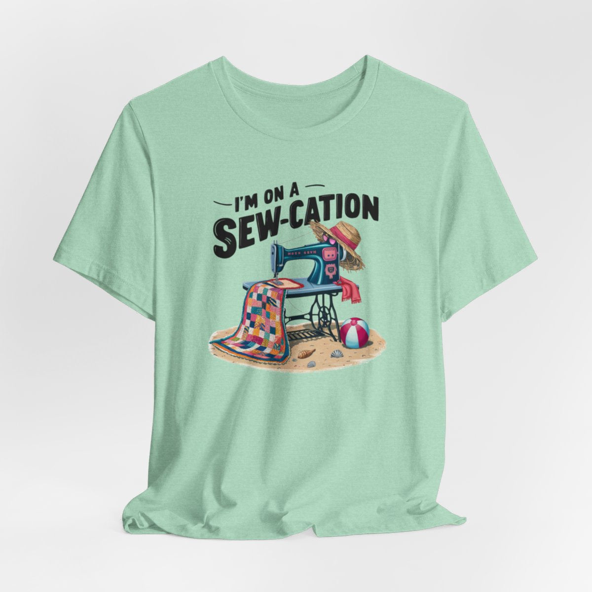 A Heather Mint fun Quilting T-shirt of a vintage sewing machine on a beach with a colorful quilt, sun hat, and beach ball. Text above reads "I'm on a Sew-cation" in bold letters.