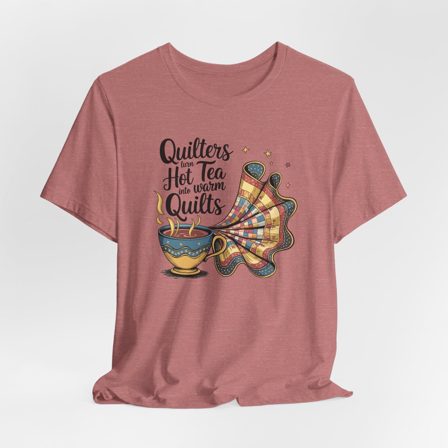 A Heather Mauve t-shirt with a whimsical design of a teacup transforming into a patchwork quilt, accompanied by the phrase 'Quilters turn hot tea into warm quilts.' Ideal for quilters and tea lovers