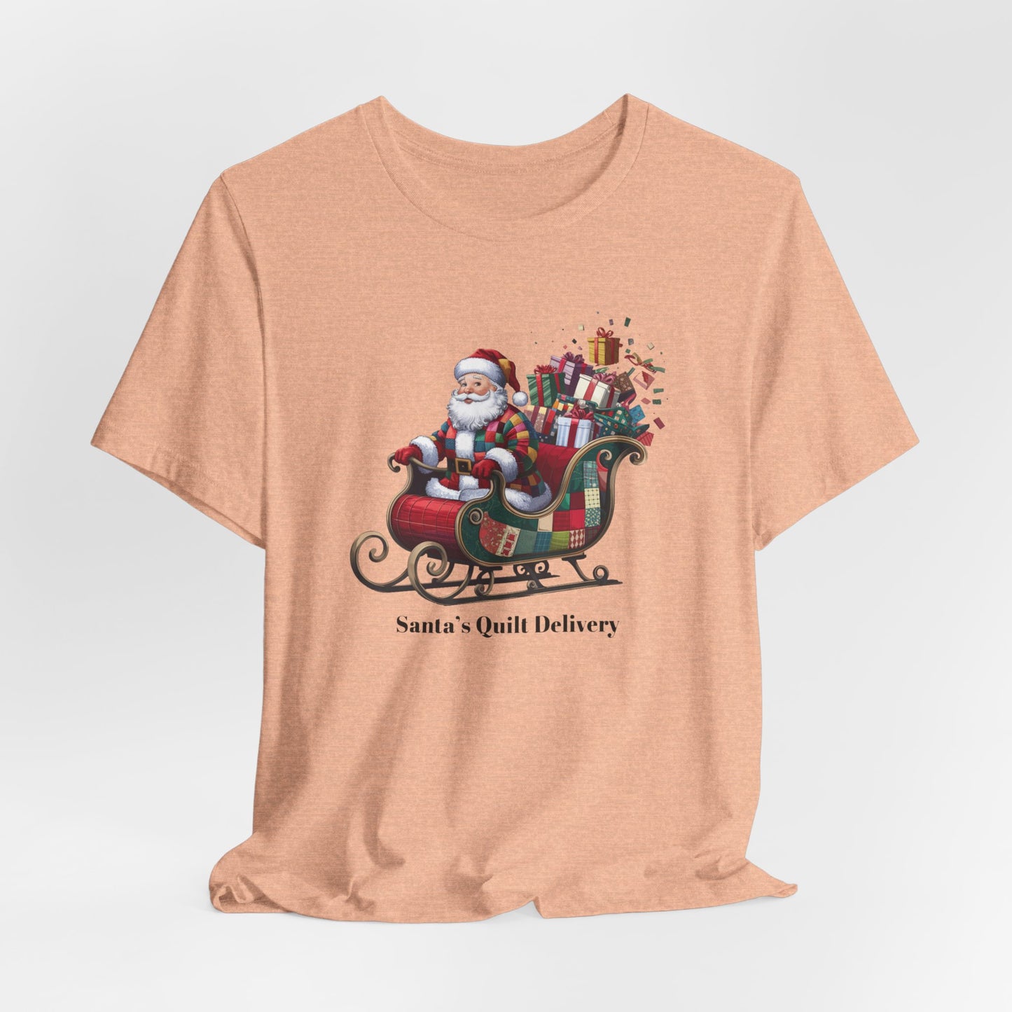 A Heather Peach christmas quilting t-shirt featuring Santa Claus wearing a patchwork quilt coat, sitting in a sleigh filled with gifts and quilts, with the phrase "Santa's Quilt Delivery" below, combining holiday cheer and quilting fun.