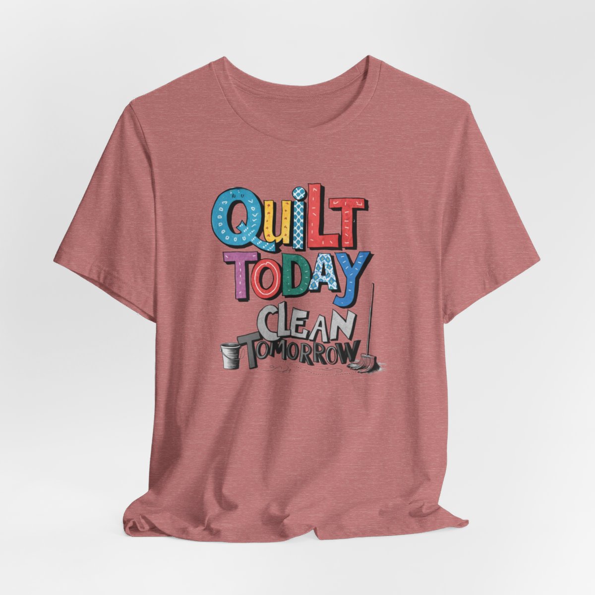 A Heather Mauve funny quilting T-Shirt with the phrase Quilt Today, Clean Tomorrow in a bold and cheerful font with a broom off to the side