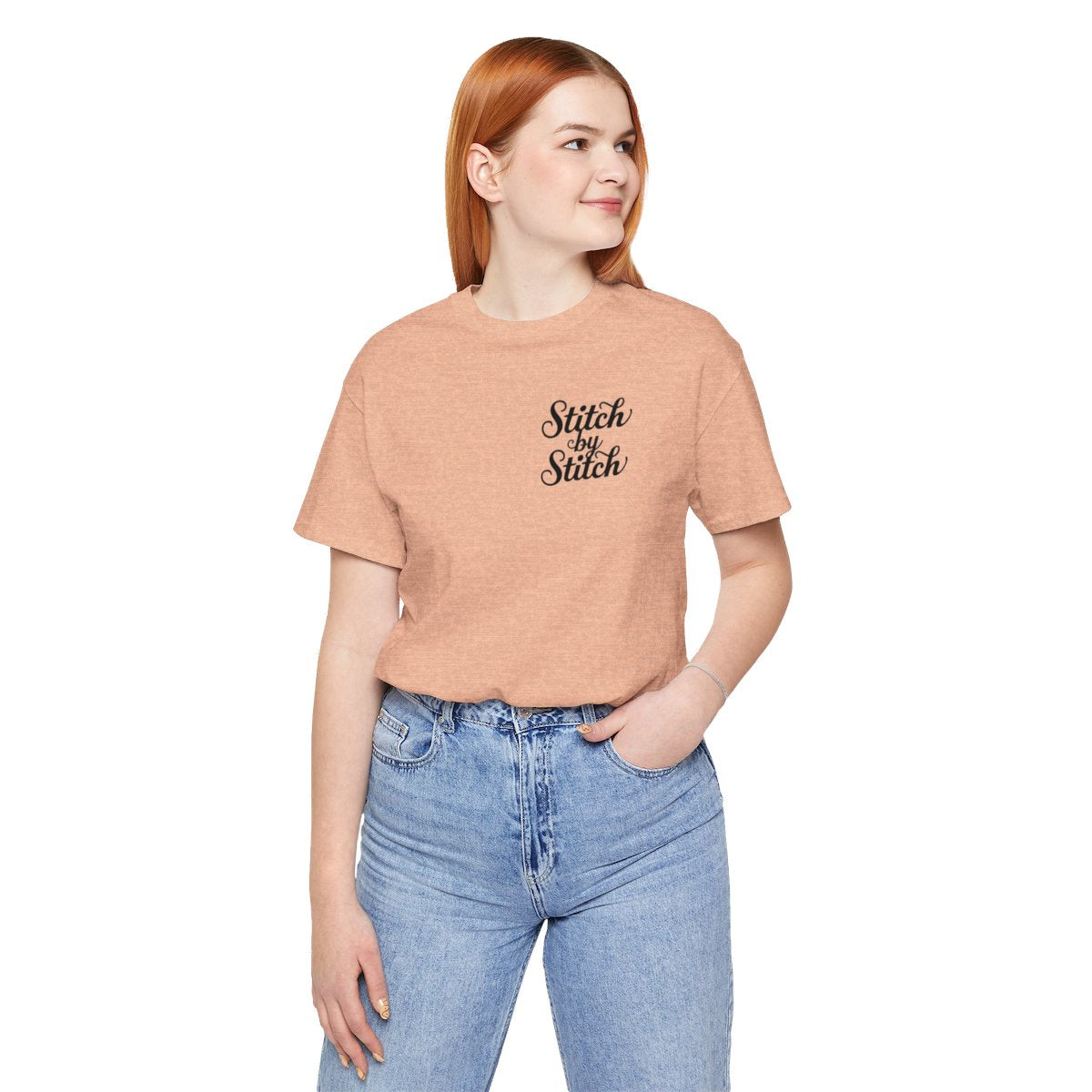 A Heather Peach T-Shirt featuring the phrase "Stitch by Stitch". The word "Stitch" is written twice in a flowing script font, while "by" is centered between them in a smaller, sans-serif font. The design has a clean, minimalist aesthetic.