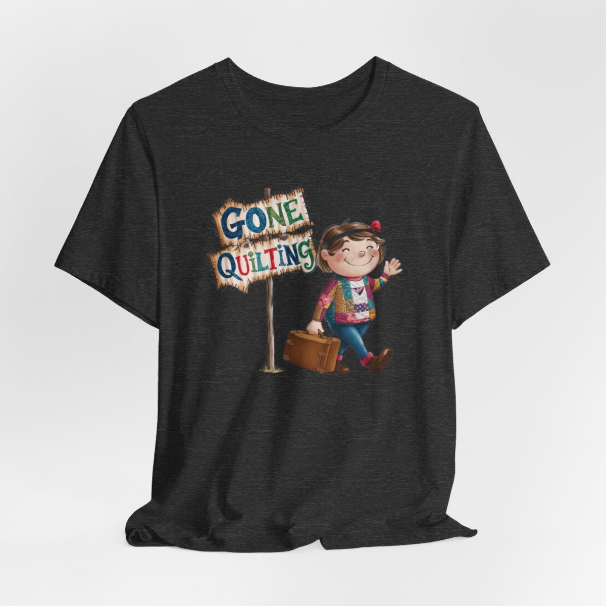 A Dark Grey Heatherfun quilting T-Shirt with a cartoon illustration of a smiling girl wearing a patchwork jacket, carrying a suitcase next to a wooden sign that reads "Gone Quilting" in colorful letters.