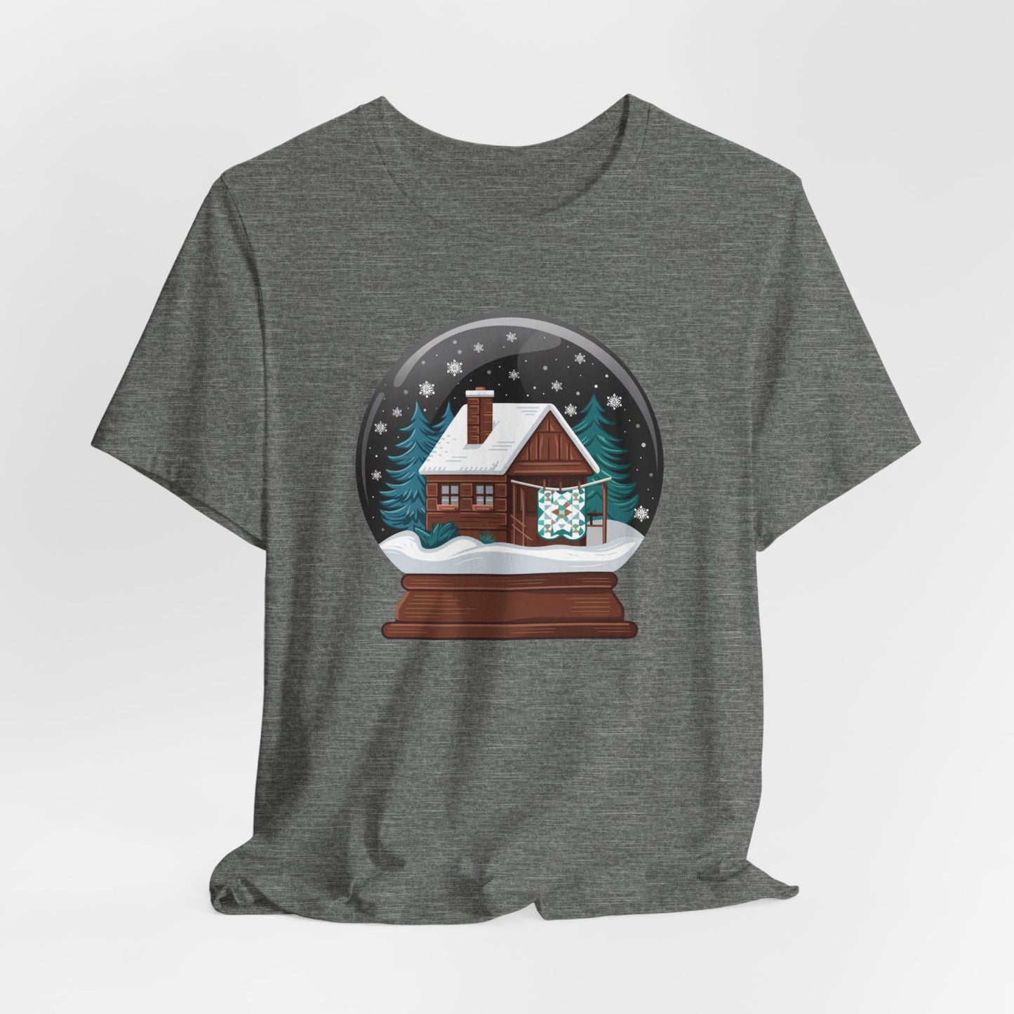 A Deep Heather christmas quilting t-shirt featuring a cozy log cabin covered in snow, with a quilt hanging outside, set inside a snow globe with swirling snowflakes, blending quilting and holiday warmth.