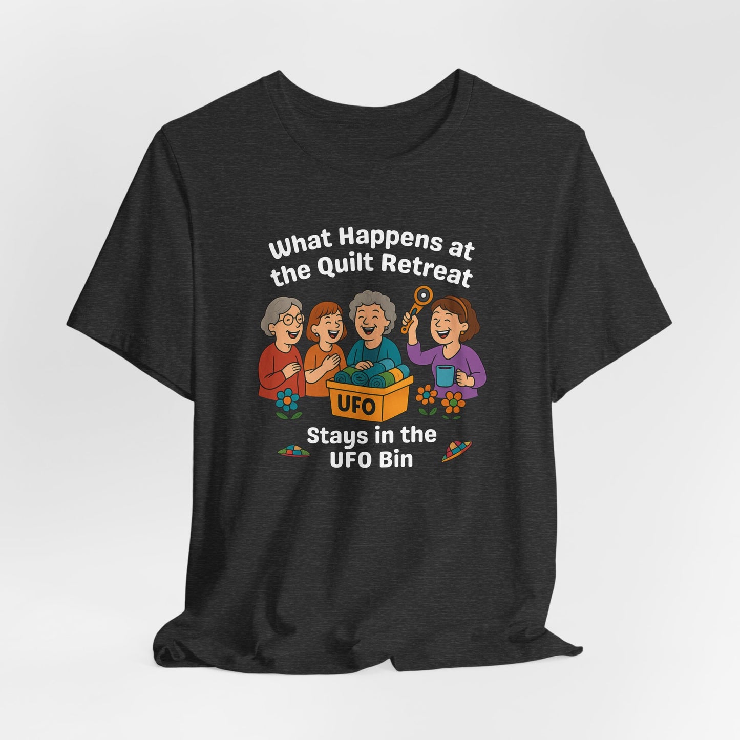A A Dark Grey Heather t-shirt featuring a cartoon design of four laughing women at a quilt retreat with a bin labeled “UFO” full of fabric. Surrounded by patchwork flying saucers and flowers. Text reads “What happens at the quilt retreat stays in the UFO bin.”