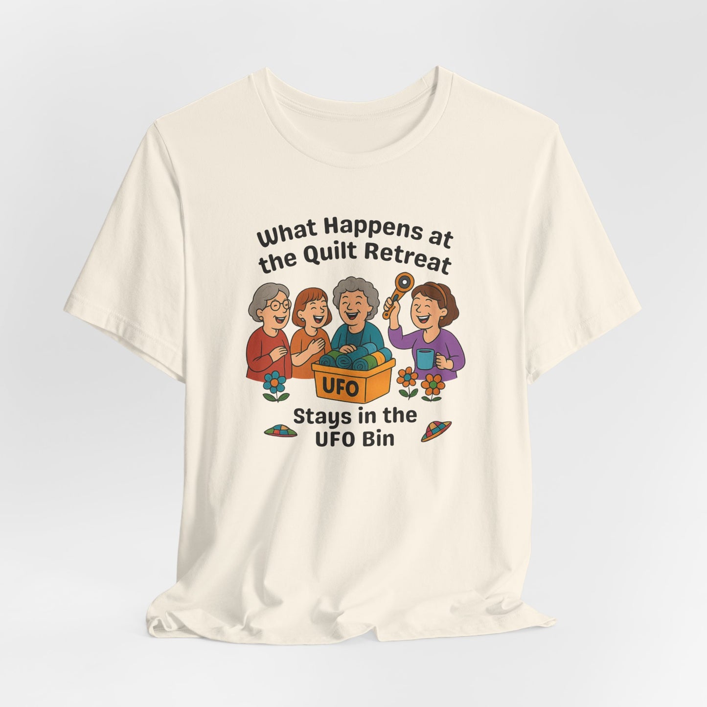 A A Natural t-shirt featuring a cartoon design of four laughing women at a quilt retreat with a bin labeled “UFO” full of fabric. Surrounded by patchwork flying saucers and flowers. Text reads “What happens at the quilt retreat stays in the UFO bin.”