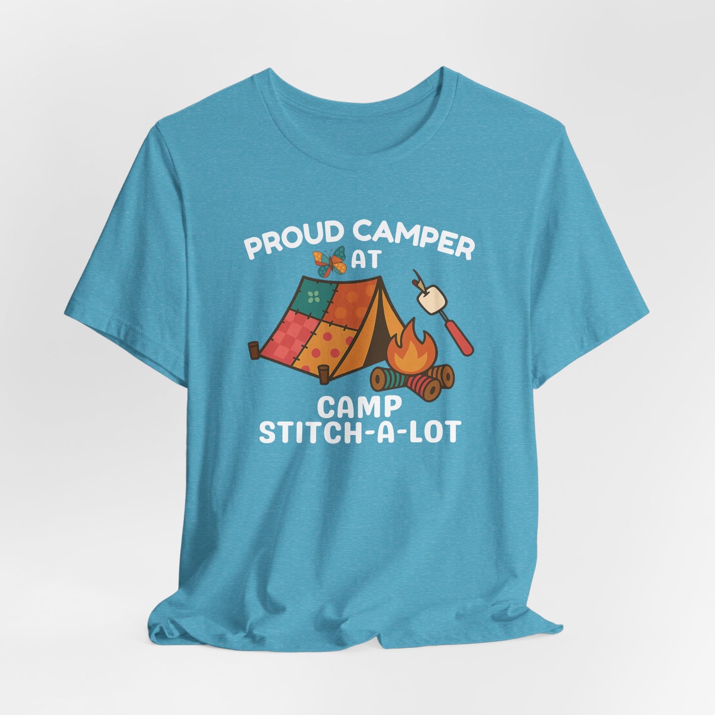 A A Heather Aqua t-shirt featuring a colorful design with a patchwork tent, thread spool campfire, marshmallow on a seam ripper stick, and a butterfly. Text reads "Proud Camper at Camp Stitch-a-Lot." Fun gift for quilting retreats.