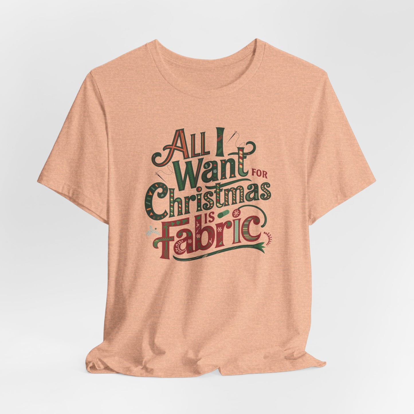 A Heather Peach festive quilting christmas T-shirt featuring the phrase "All I Want for Christmas is Fabric," with fun sewing and quilting accents like needles and threads swirling around the text.