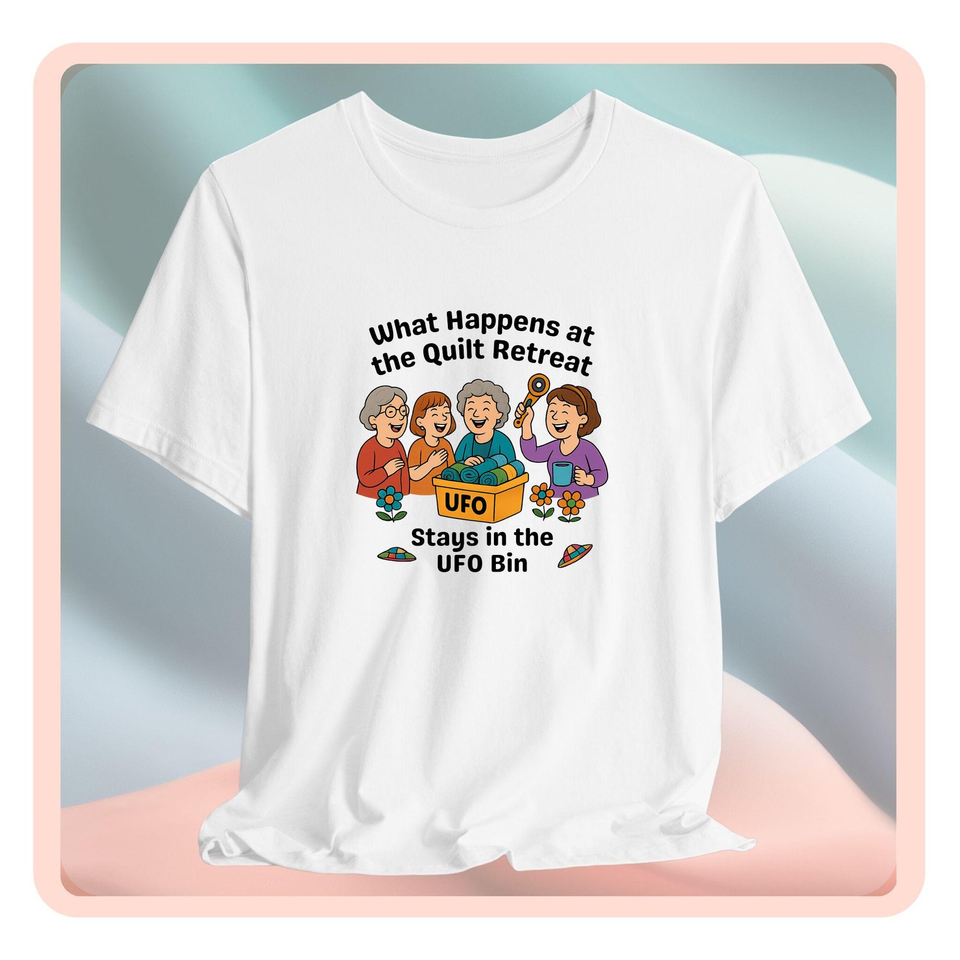 A A t-shirt featuring a cartoon design of four laughing women at a quilt retreat with a bin labeled “UFO” full of fabric. Surrounded by patchwork flying saucers and flowers. Text reads “What happens at the quilt retreat stays in the UFO bin.”