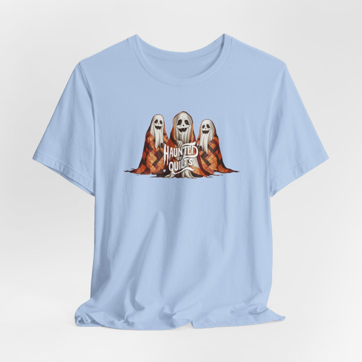 A Baby Blue T-Shirt of three smiling cartoon ghosts covered in colorful patchwork quilts. The quilts feature orange, brown, and black patterns. Text below reads "HAUNTED BY QUILTS" in a spooky white font.