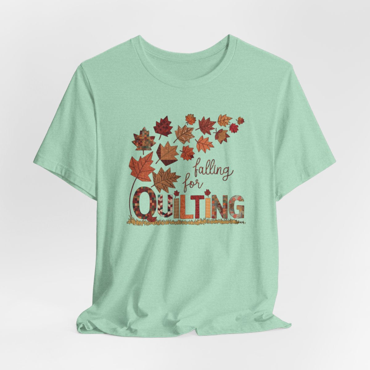 A Heather Mint thanksgiving quilting t-shirt featuring autumn leaves with various quilting patterns inside. Text reads "falling for QUILTING" with letters in patchwork style. Warm fall colors and quilting motifs combine in a seasonal design.