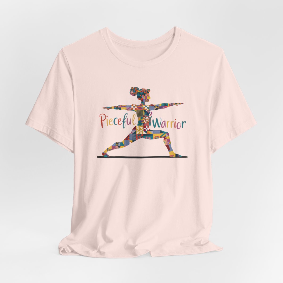 A Soft Pink Tee featuring a colorful patchwork silhouette of woman in yoga warrior pose. Text reads "Pieceful Warrior" in multicolored lettering.