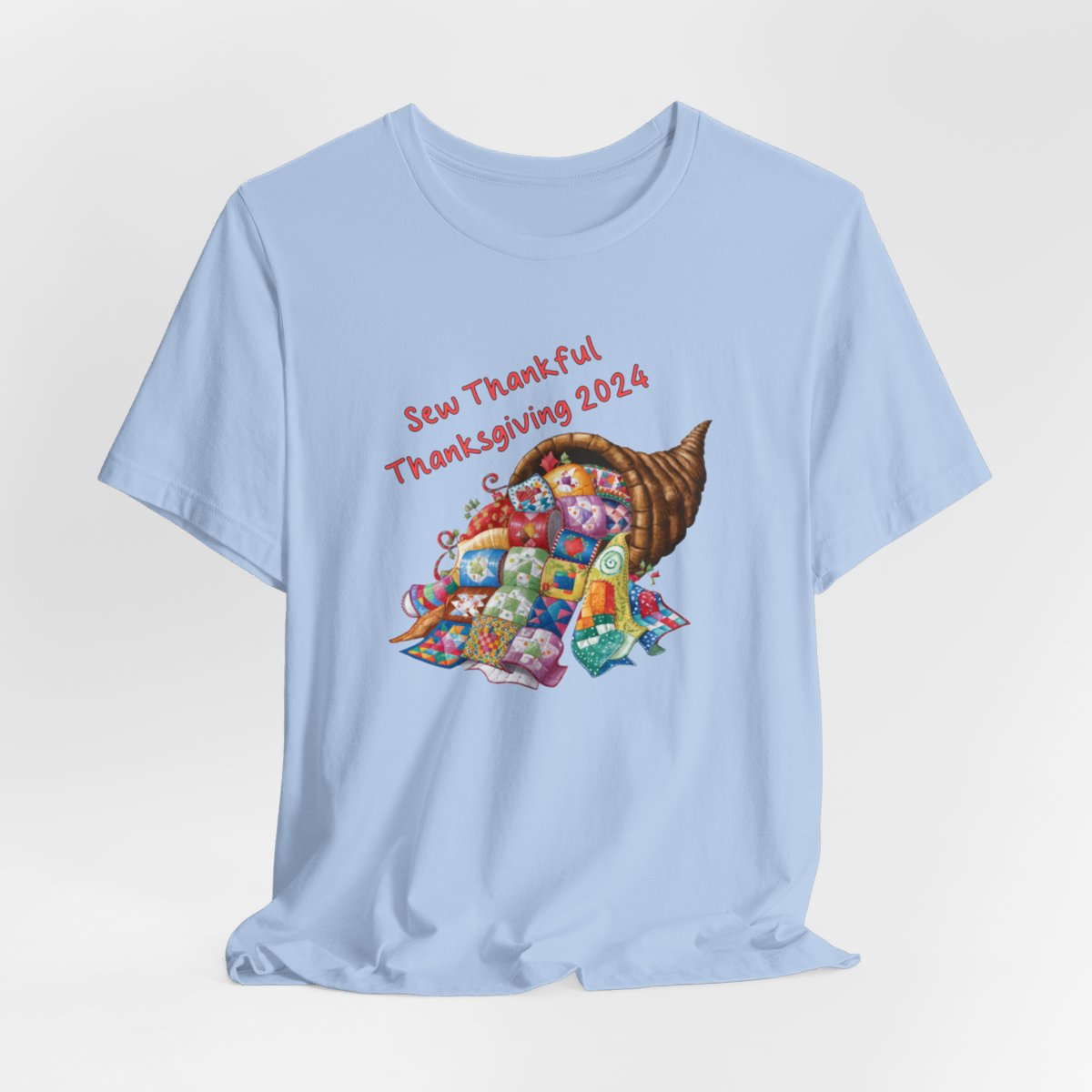A Baby Blue thanksgiving quilting t-shirt featuring a colorful quilted cornucopia overflowing with various quilt patterns and designs. Text reads "Sew Thankful Thanksgiving 2024" in red. Vibrant illustration combines quilting motifs with Thanksgiving imagery.