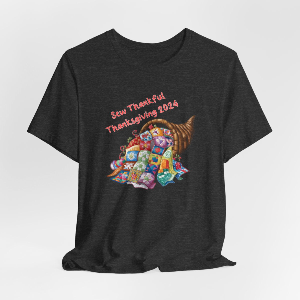 A Dark Grey Heather thanksgiving quilting t-shirt featuring a colorful quilted cornucopia overflowing with various quilt patterns and designs. Text reads "Sew Thankful Thanksgiving 2024" in red. Vibrant illustration combines quilting motifs with Thanksgiving imagery.