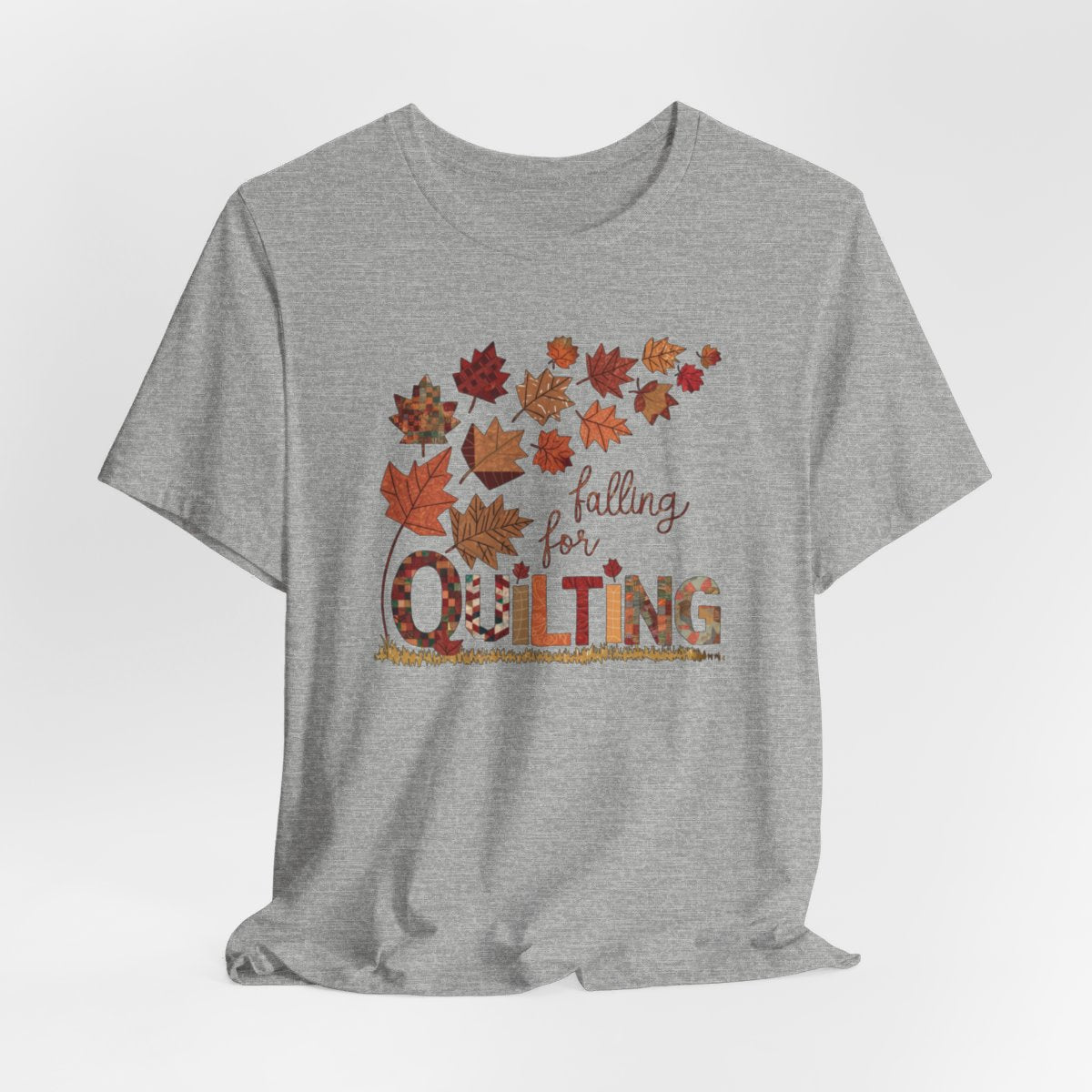 An Athletic Heather thanksgiving quilting t-shirt featuring autumn leaves with various quilting patterns inside. Text reads "falling for QUILTING" with letters in patchwork style. Warm fall colors and quilting motifs combine in a seasonal design.