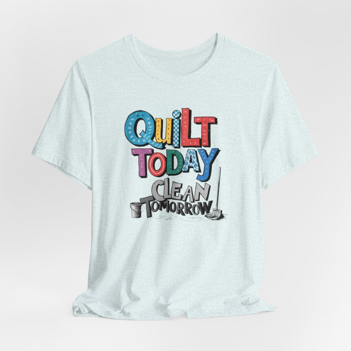 A Heather Ice Blue funny quilting T-Shirt with the phrase Quilt Today, Clean Tomorrow in a bold and cheerful font with a broom off to the side