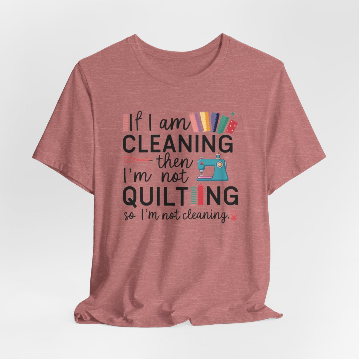 A Heather Mauve funny quilting T-Shirt with the phrase If I am Cleaning then I'm not Quilting in a bold and cheerful font with images of quilting icons and supplies