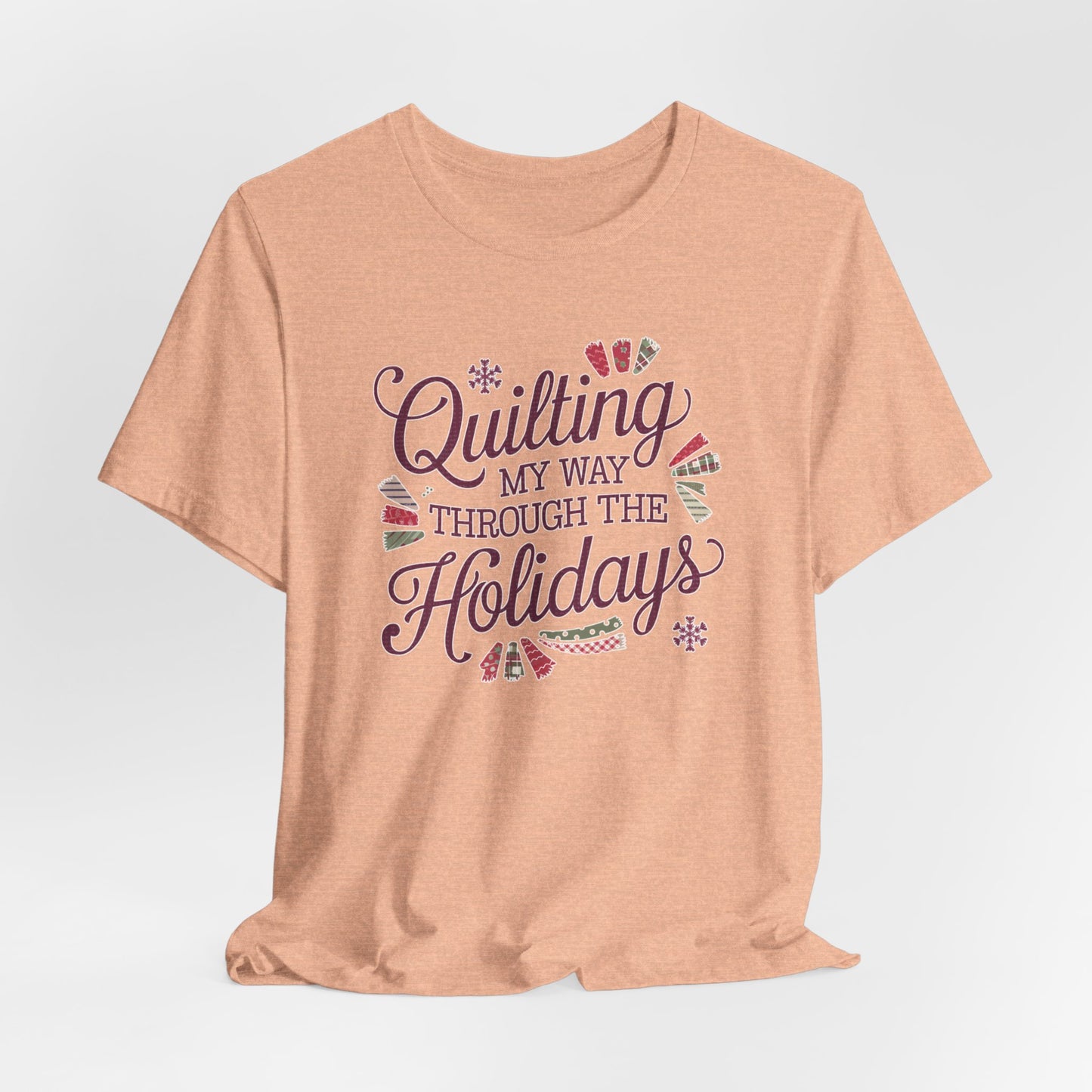 A Heather Peach playful holiday-themed t-shirt with the phrase "Quilting My Way Through the Holidays" surrounded by quilting-inspired decorations like fabric swatches and snowflakes.