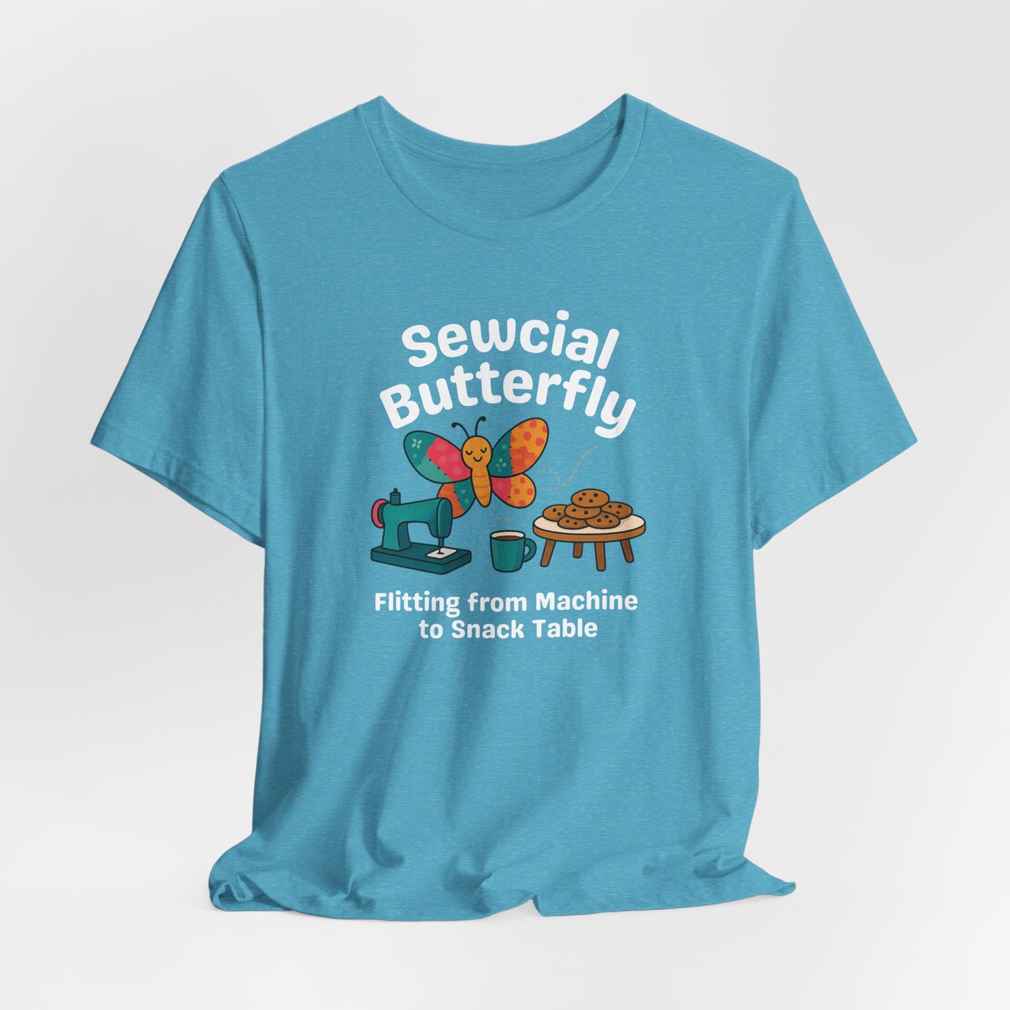 A A Heather Aqua t-shirt with a colorful design showing a patchwork butterfly flying between a sewing machine and a table of cookies. Text reads “Sewcial Butterfly – Flitting from Machine to Snack Table.” Funny gift for quilters