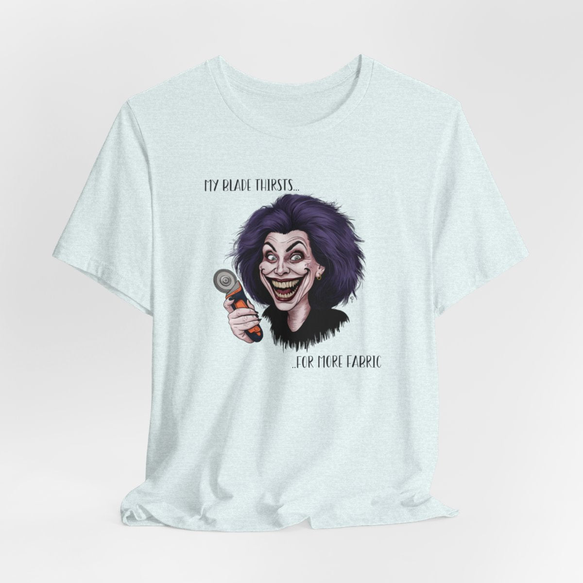 A Heather Ice Blue T-shirt showing a cartoonish character with wild purple hair and an exaggerated grin, holding a rotary cutter. Text reads "My blade thirsts... for more fabric", humorously depicting a quilter's endless desire for new materials.