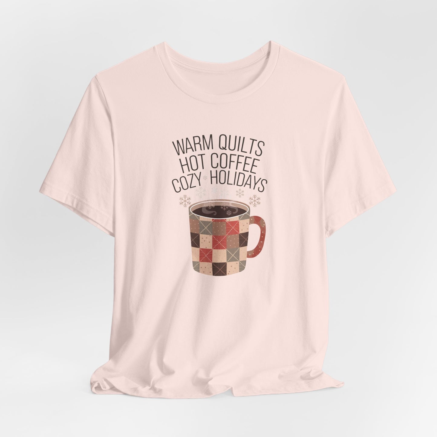 A Soft Pink christmas quilting t-shirt featuring a cozy patchwork quilt-themed coffee mug, surrounded by snowflakes, with the phrase "Warm Quilts, Hot Coffee, Cozy Holidays" above the mug, celebrating the warmth of quilts and coffee during the holiday season.