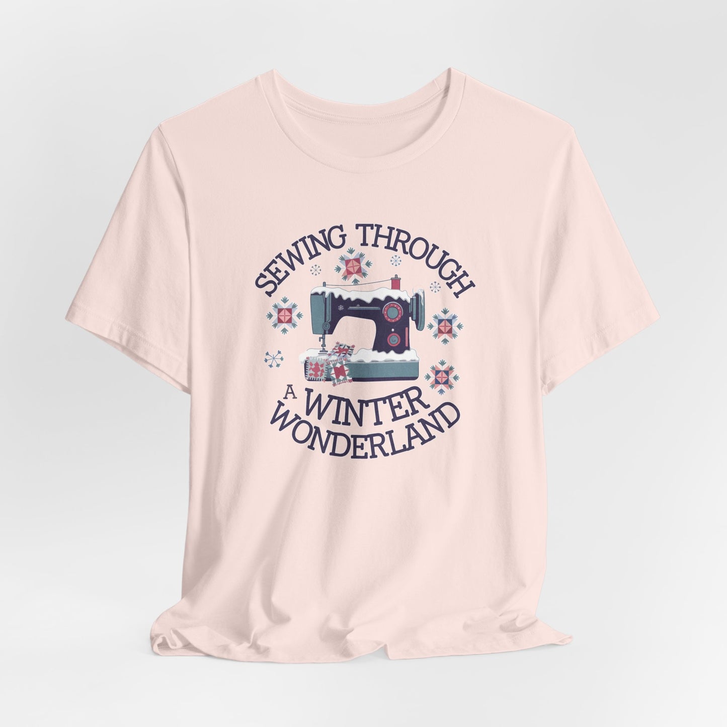A Soft Pink winter quilting t-shirt featuring a sewing machine covered in snow, surrounded by quilted snowflakes, with the phrase "Sewing Through a Winter Wonderland" above and below the image.